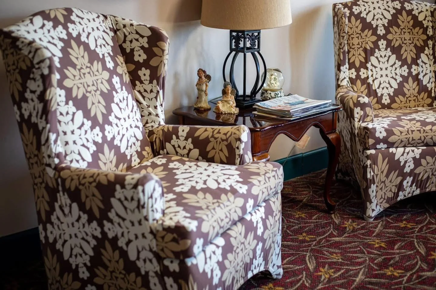 A cozy living room corner with floral patterned armchairs, a wooden side table, a table lamp, and decorative figurines, with a patterned carpet on the floor.