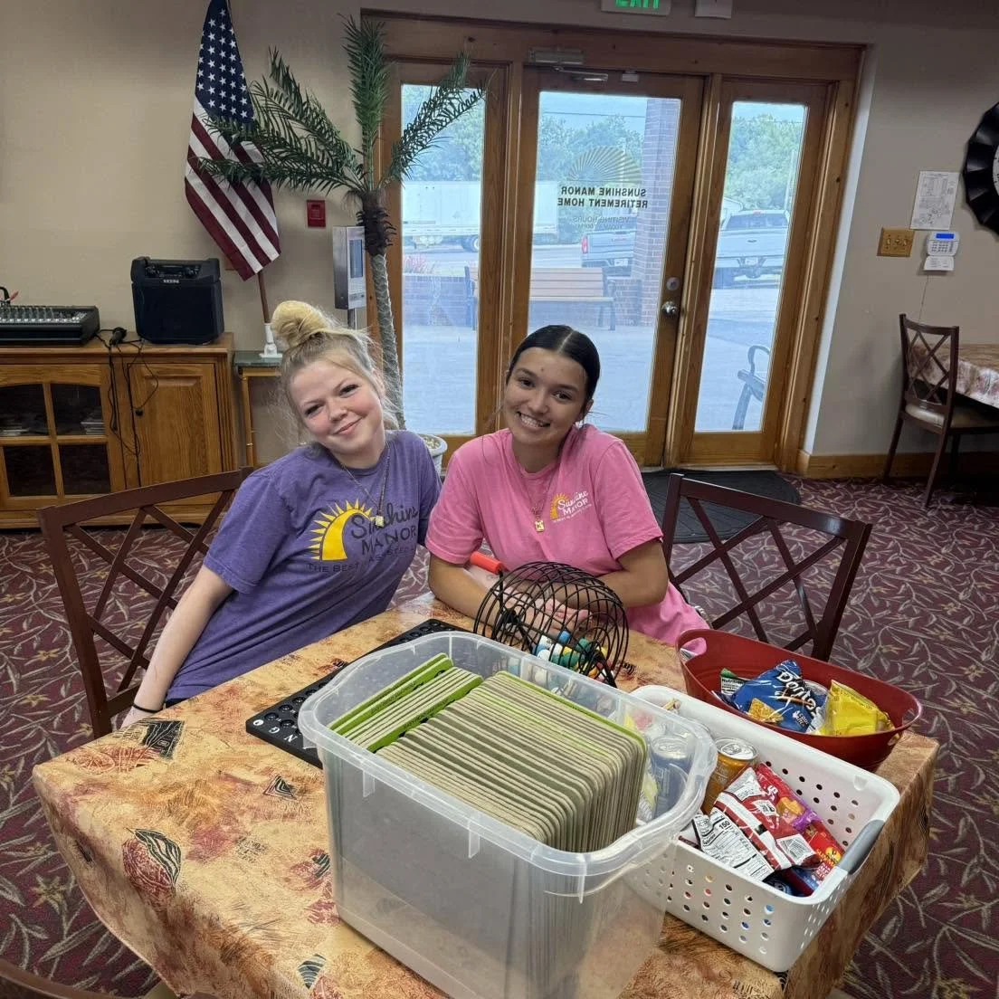 Two girls sitting at a table smiling, surrounded by snacks, with a large potted plant and a flag behind them. They are inside a room with a glass door and windows showing an outdoor parking lot.