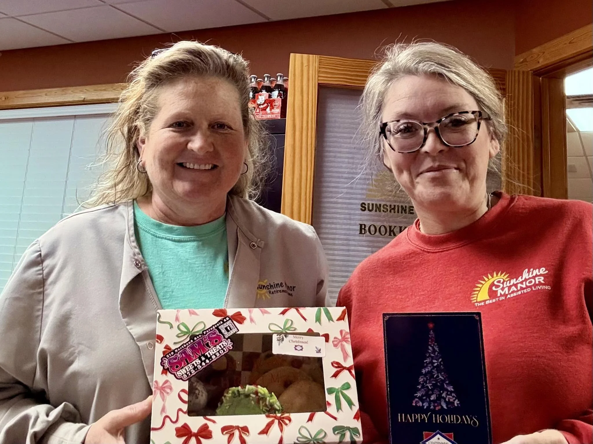 Two women standing indoors, smiling at the camera. One is holding a holiday-themed box. The woman on the left has curly hair and is wearing a light-colored jacket, while the woman on the right has glasses and short gray hair, wearing a red shirt. Behind them is a wooden door frame, a window with blinds, and a sign that reads 'SUNSHINE MANOR.'