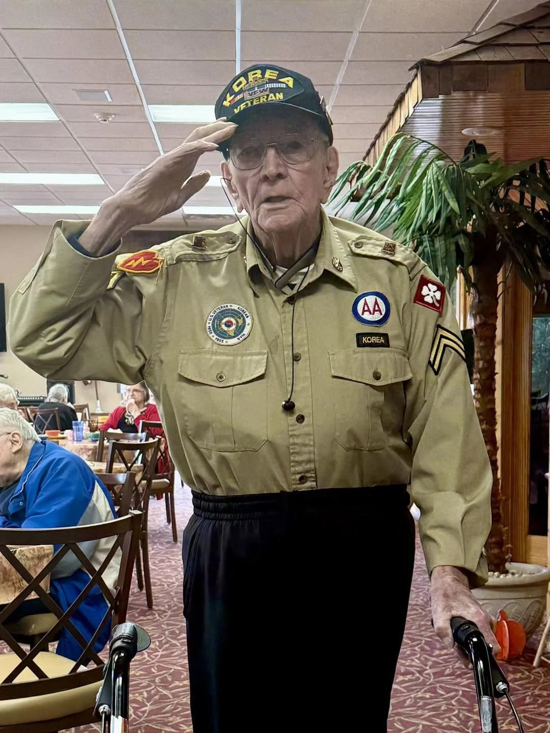 An elderly man wearing a military uniform and a cap with 'KOREA VETERAN' salutes inside a restaurant, holding a walker in one hand.