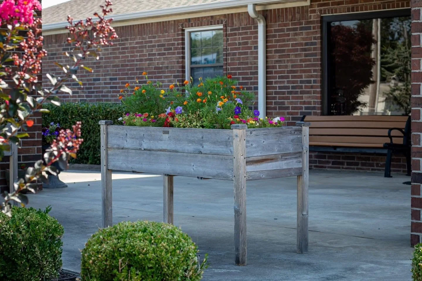 Wooden planter box filled with colorful flowers on a concrete porch in front of a brick house with window and sliding glass door.