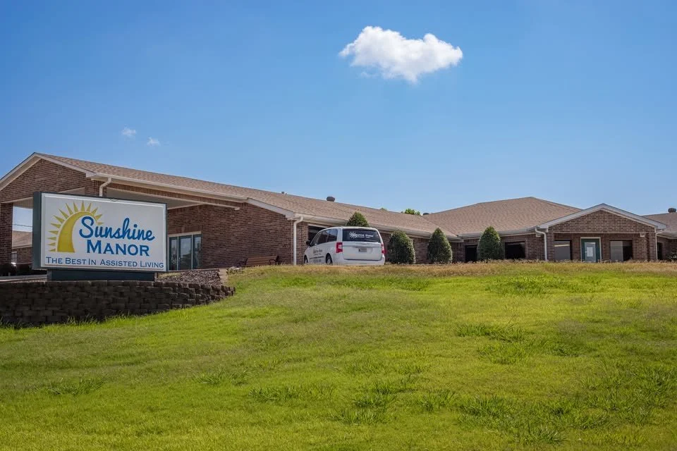 Sunshine Manor assisted living facility with a brick building, a parking area with a white van, and a well-kept grassy lawn, under a blue sky with a few clouds.