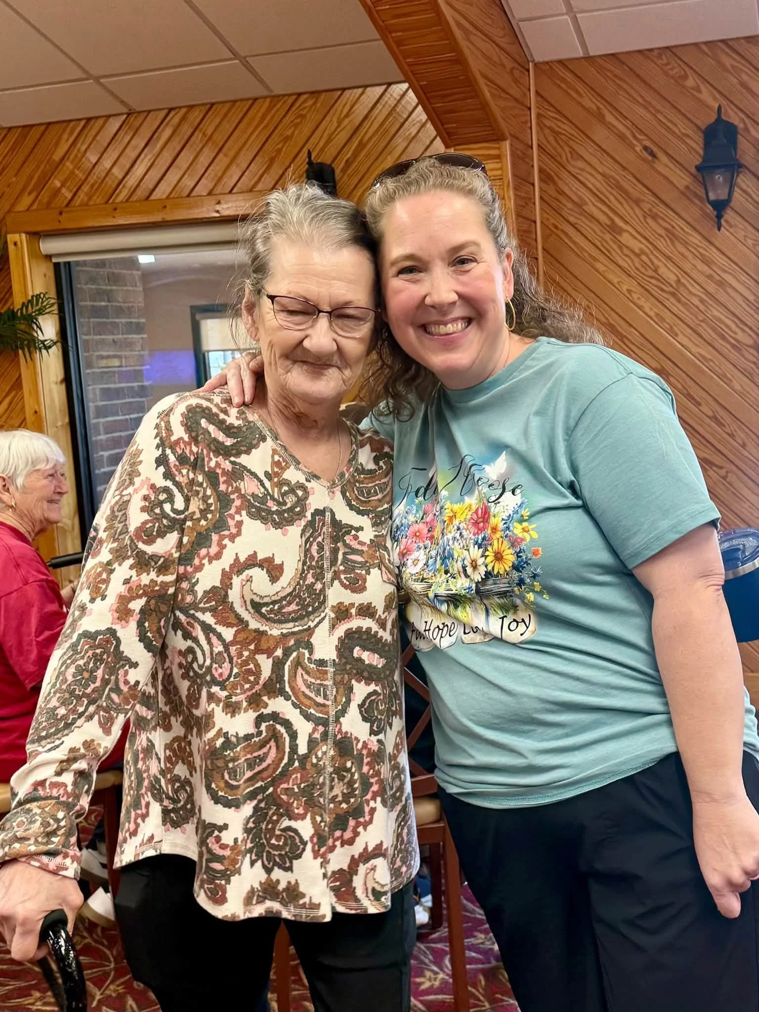Two women smiling and hugging each other, one elderly and the other middle-aged, inside a wooden-paneled room with a window and brick wall visible in the background.