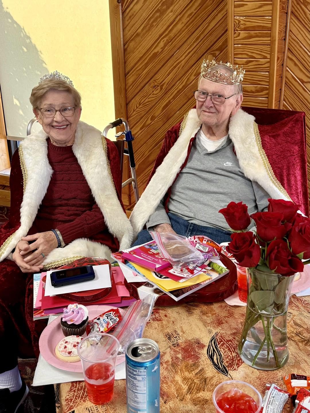 An elderly couple sitting at a table, dressed in festive attire with crowns and capes, celebrating with Valentine's Day decorations, desserts, and drinks.
