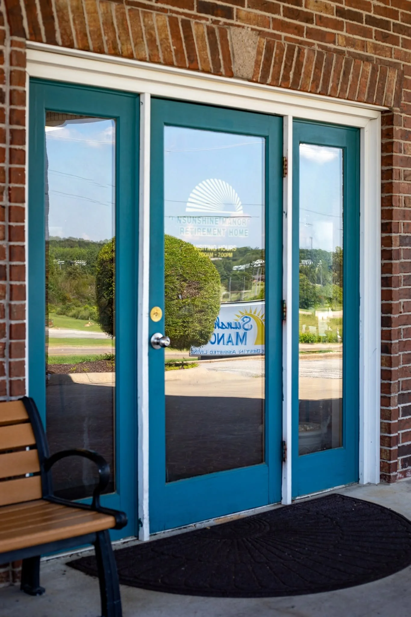 Glass door with a blue frame reflecting trees and a blue sky, with a sign for Sunshine Manor Retirement Home and operating hours, brick wall, wooden bench nearby, and green landscape outside.