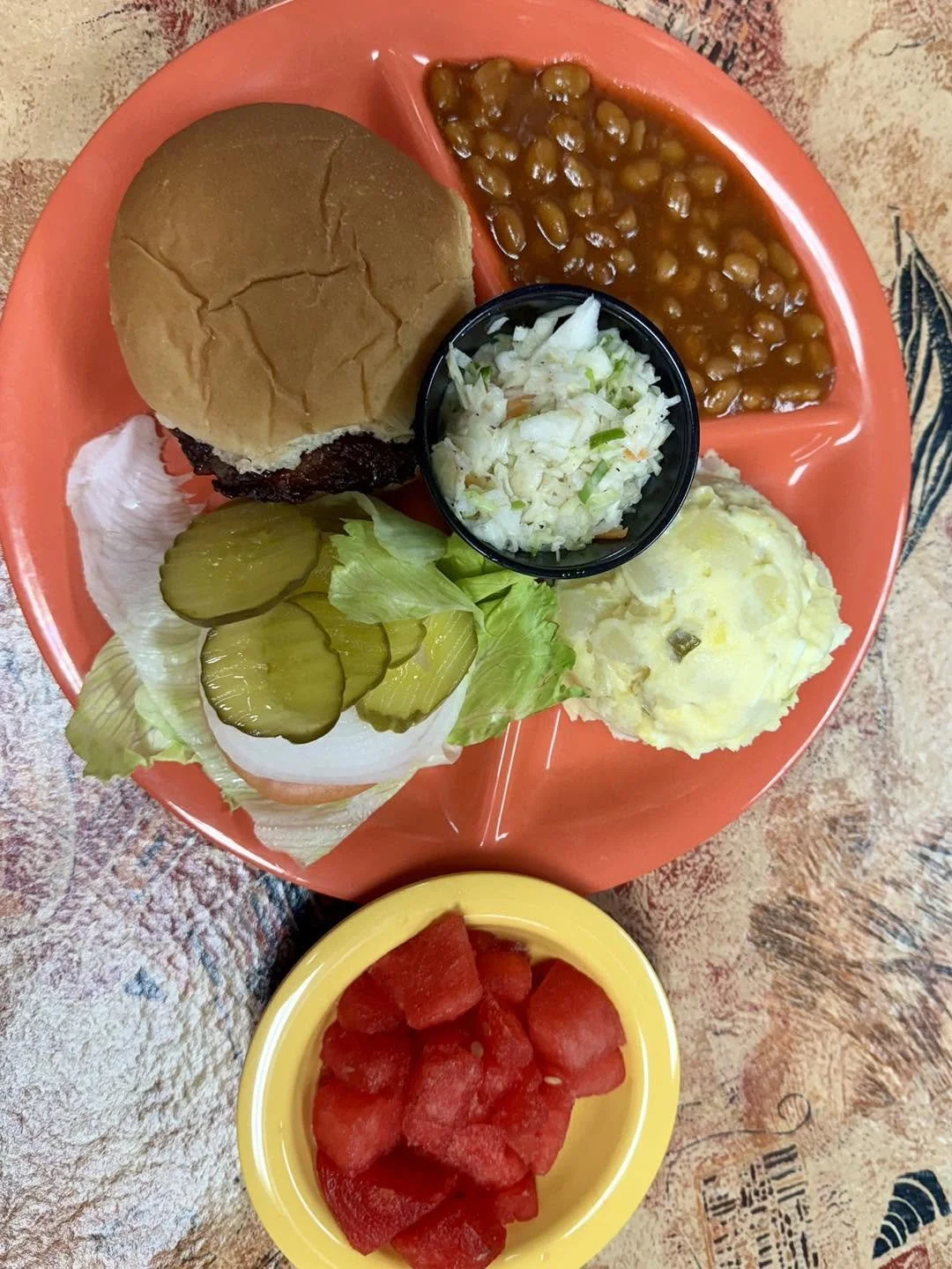 A meal served on a divided pink plate with a small yellow bowl of watermelon, consisting of a hamburger bun with a beef patty, baked beans, coleslaw, potato salad, and sliced pickles on lettuce.