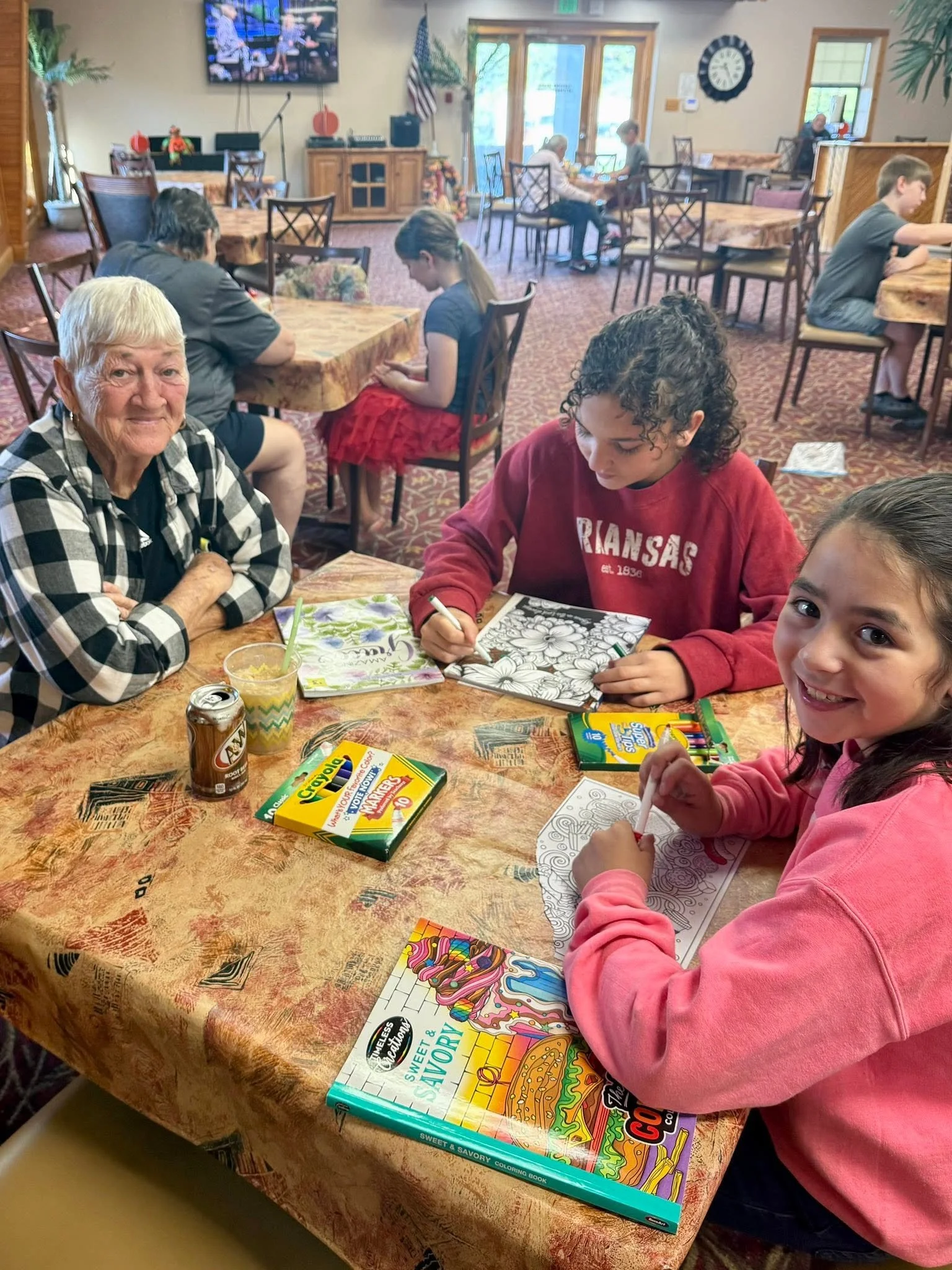 A group of children and an elderly woman sitting at a table in a restaurant, coloring in coloring books. The table has coloring supplies, snacks, and drinks. The background shows more tables with children and adults, TV screens, and decorations.
