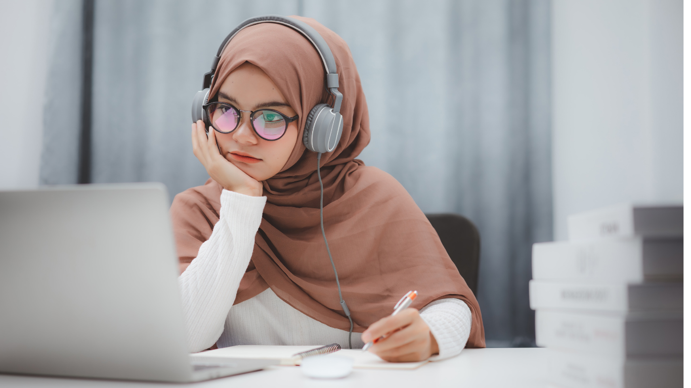 A young woman wearing a hijab and glasses is sitting at a table, looking at a laptop screen with a focused expression. She is wearing large headphones and holding a pen, with a stack of books on her right.