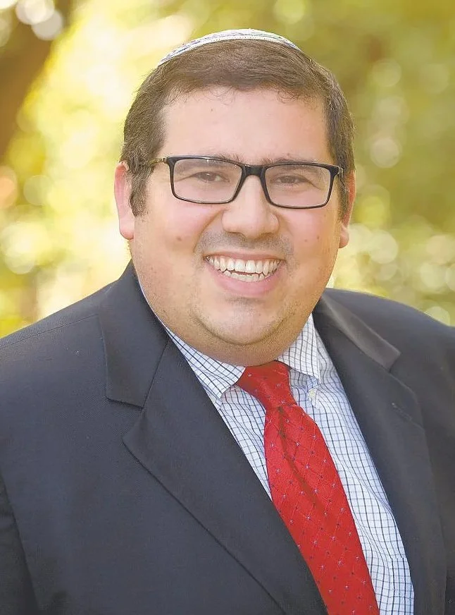 A man with short dark hair, glasses, and a big smile, wearing a dark suit, a white checkered shirt, and a red tie, standing outdoors with blurred green and yellow trees in the background.