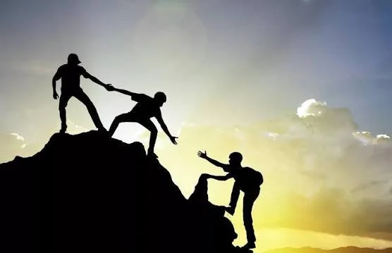 Silhouettes of three people helping each other climb a mountain during sunset.
