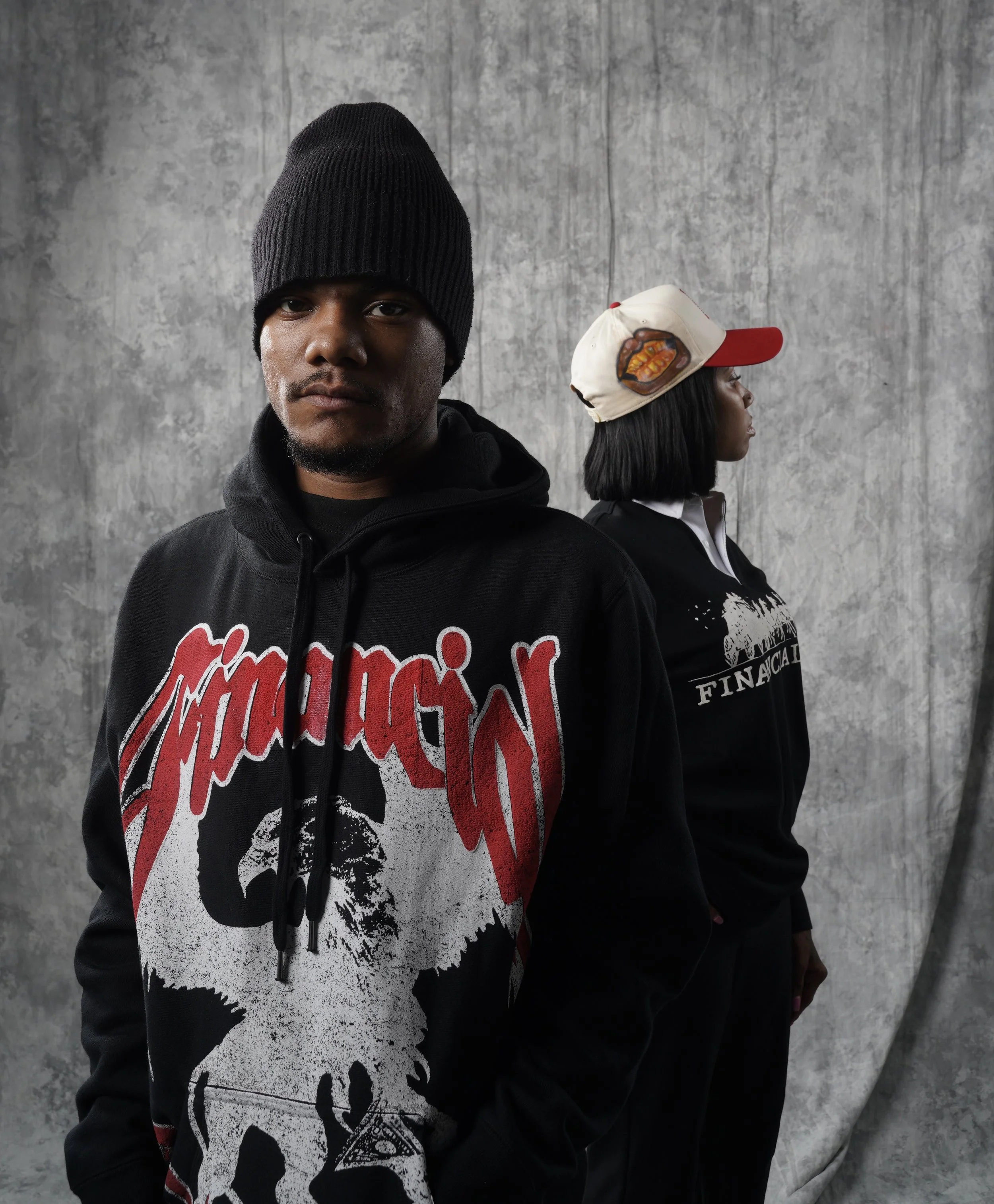 A young man wearing a black hoodie with red and white text and a black beanie, standing in front of a gray textured wall.