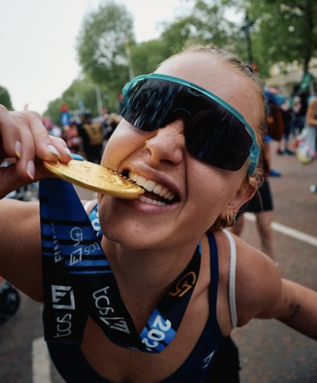 Lachende hardloopster met zonnebril bij de finish terwijl ze in een gouden medaille bijt.