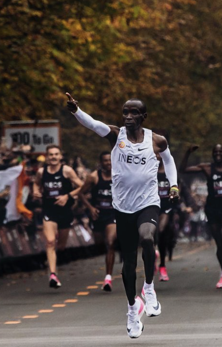 Eliud Kipchoge rent tijdens een wegwedstrijd in een wit INEOS-shirt, met hardlopers op de achtergrond.