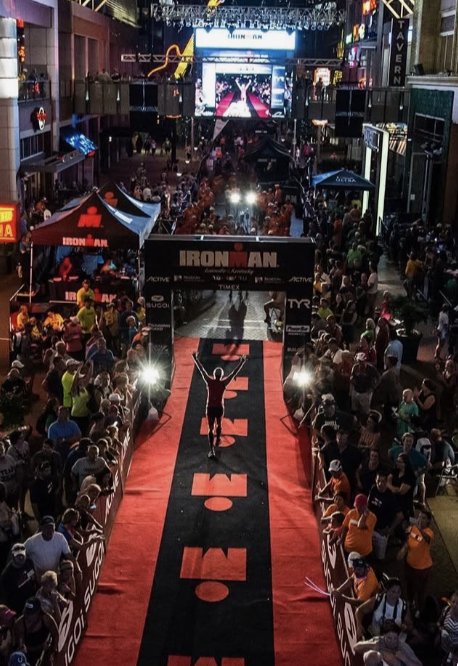 Triatleet finisht de IRONMAN-wedstrijd op een rode finishmat, omringd door een juichende menigte in een verlichte stadsstraat.