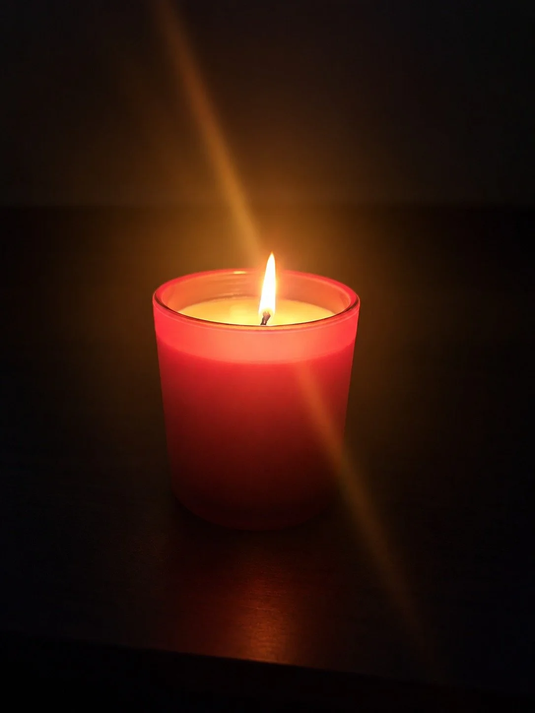 A lit red candle with a visible flame on a dark surface, casting a warm glow.