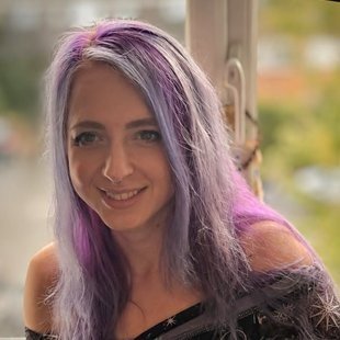 A woman with lavender and silver hair smiling indoors with a window in the background.