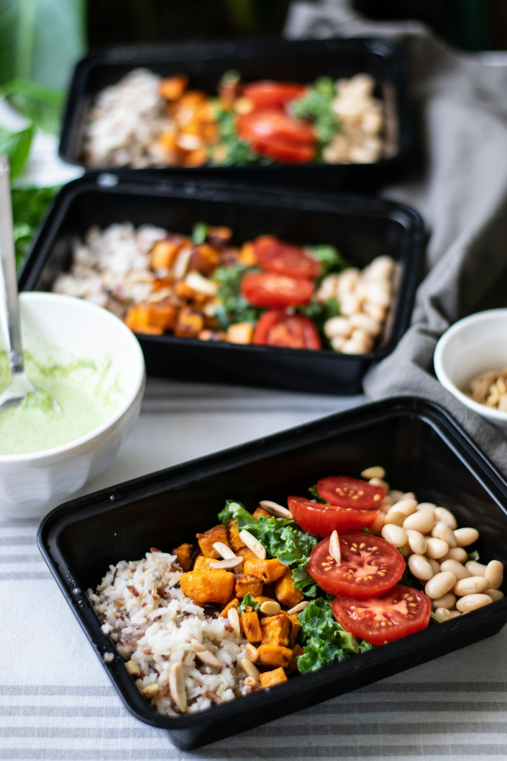 Three black meal prep containers filled with rice, cherry tomatoes, white beans, roasted sweet potatoes, and greens, on a gray and white striped table.