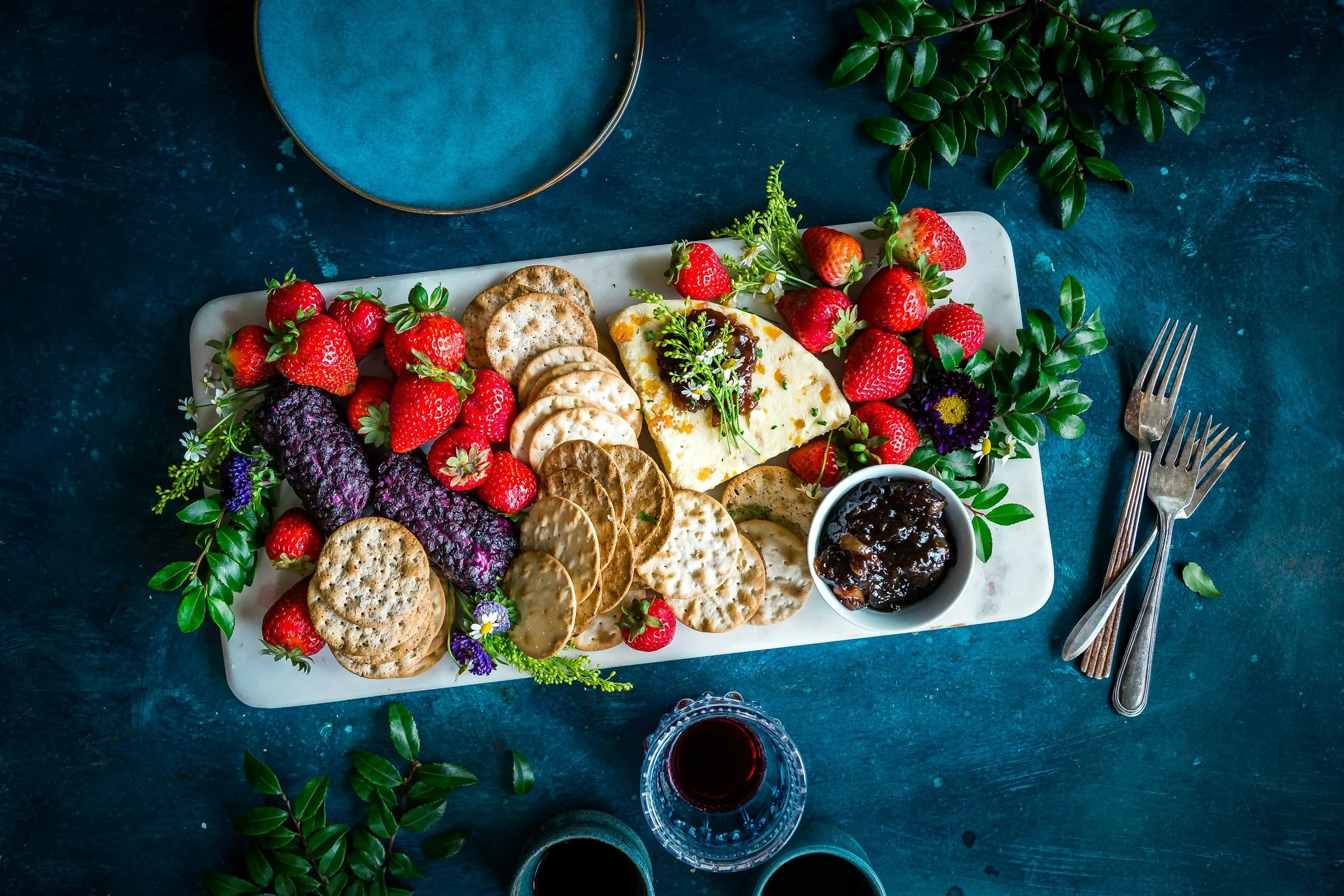 A white rectangular serving platter with assorted crackers, fresh strawberries, a wedge of cheese, purple and green herbs, and a small bowl of dark jam. There is a blue plate, floral decorations, green leaves, and two glasses of red wine on a dark blue textured surface, with two forks placed on the right side of the platter.