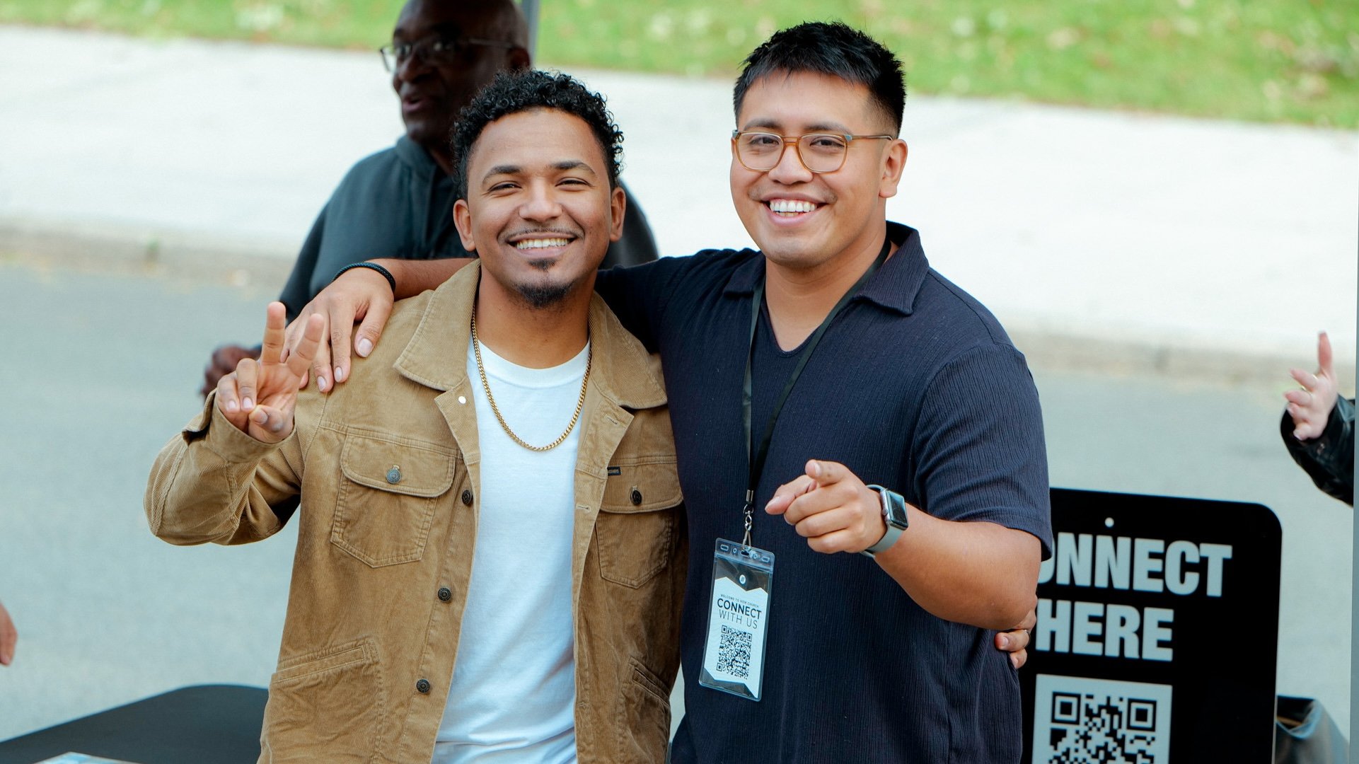 Two smiling men standing side by side at an outdoor event, with one showing a peace sign and the other pointing, behind a sign that says 'Connect Here' with a QR code, and a third person partially visible in the background.
