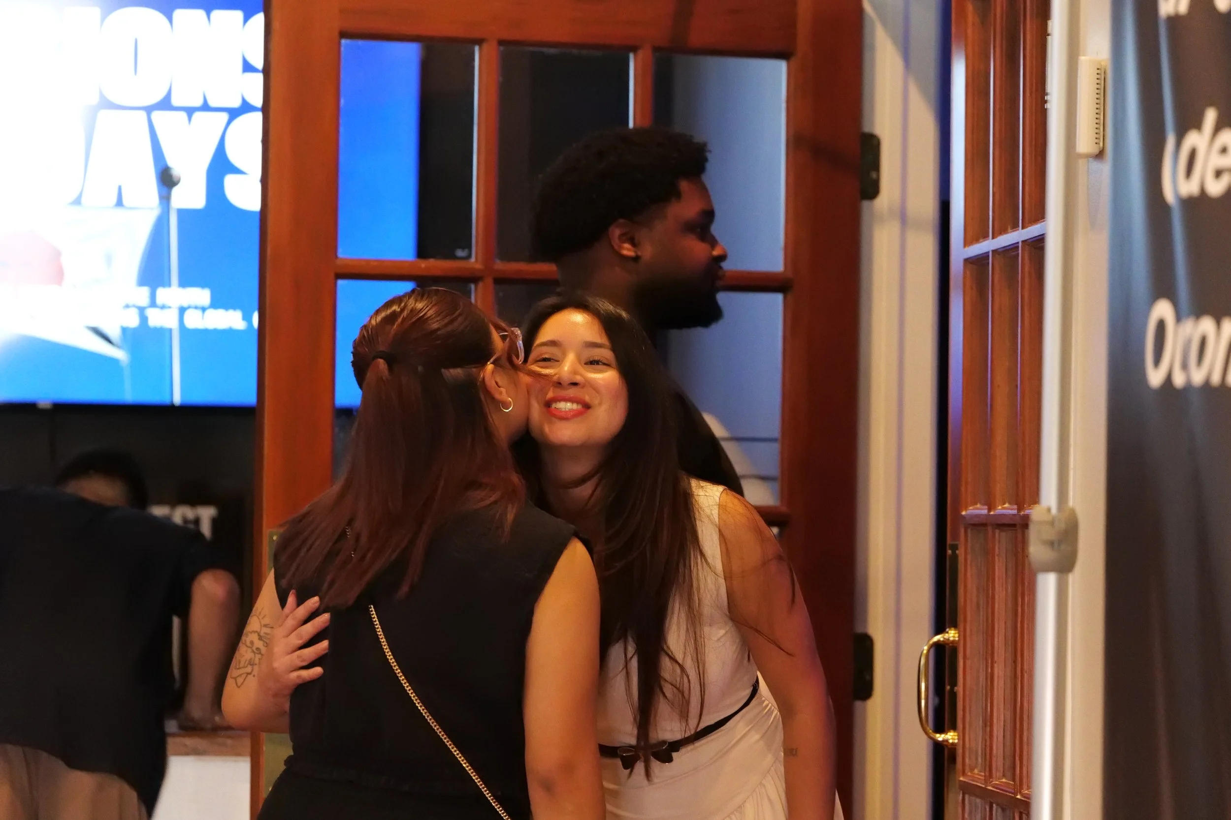 Four people, three women and one man, are inside a restaurant or bar, embracing and smiling. One woman is kissing another woman on the cheek, both smiling happily. The man is standing behind two women and looking to the right. The background shows wooden walls and a large blue sign.