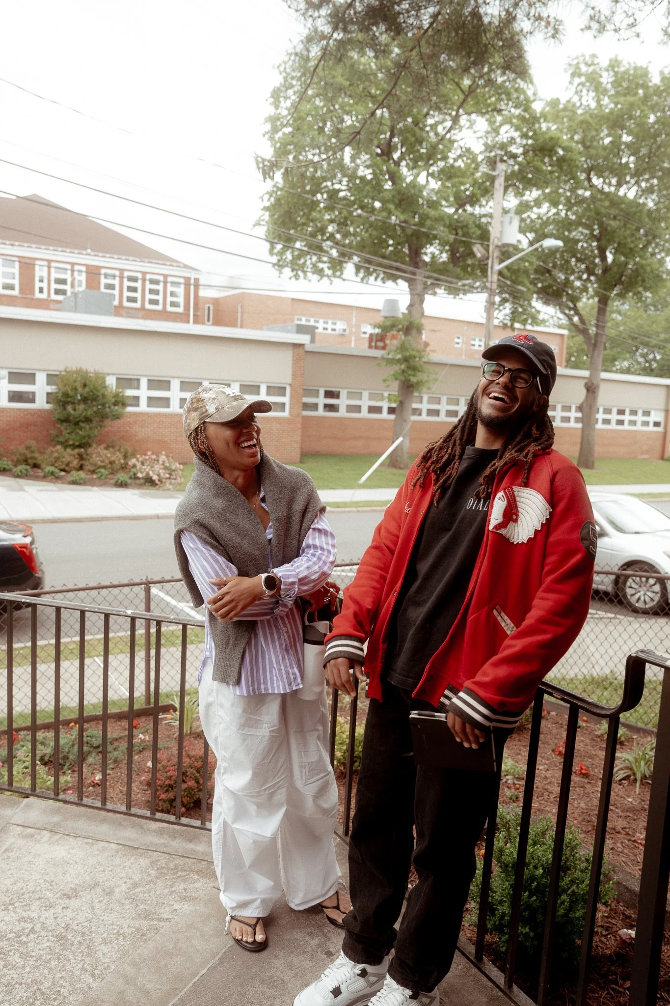 Two people, a woman and a man, standing outside on a sidewalk near a black metal fence. They are smiling and laughing. The woman is wearing a beige cap, a gray sweater over a striped shirt, and white pants. The man is wearing glasses, a black cap, a red jacket with a logo, and black pants. There are trees and buildings in the background.