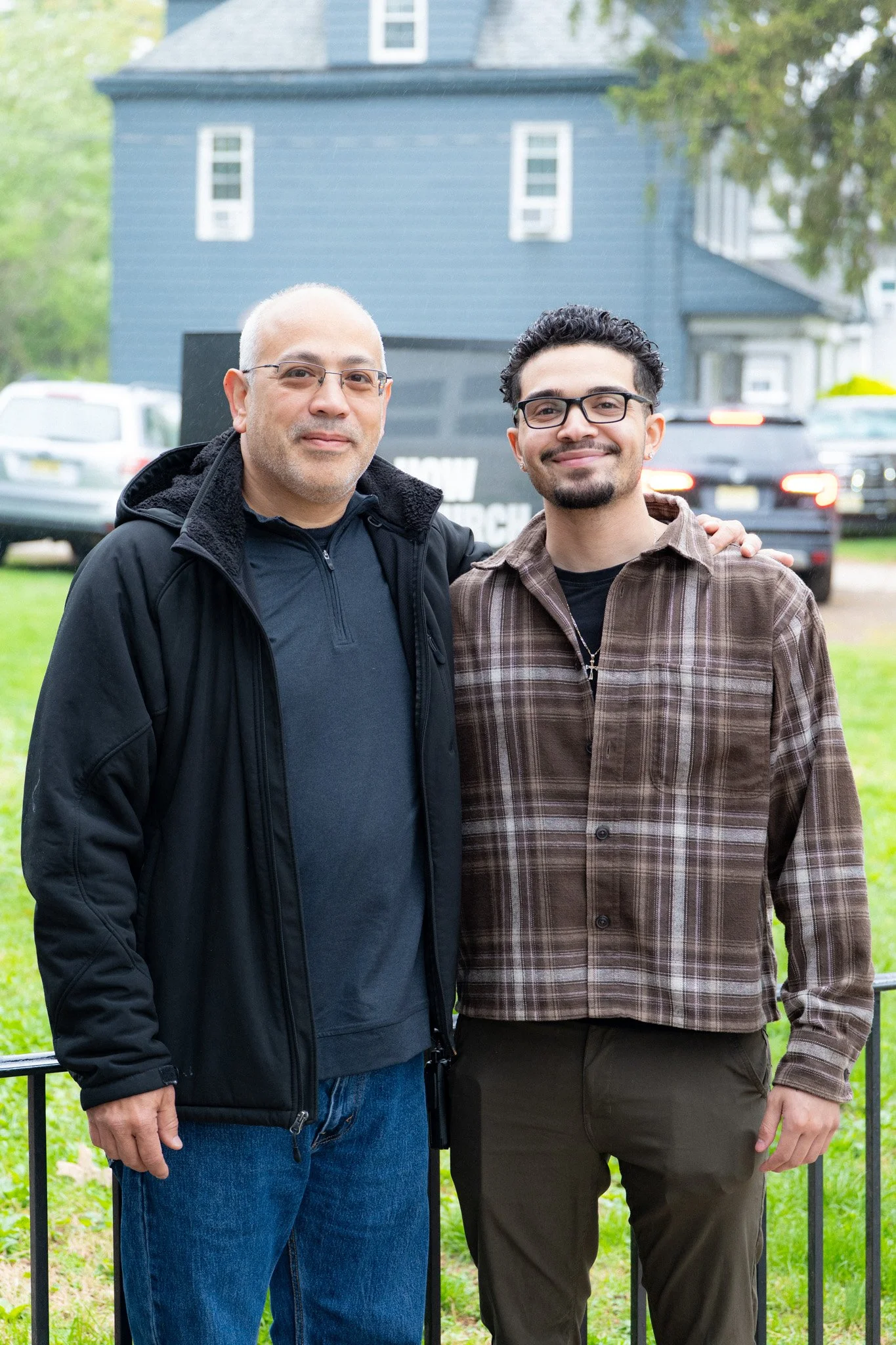 Two men outdoors standing close together, smiling, with a blue house and parked cars in the background.