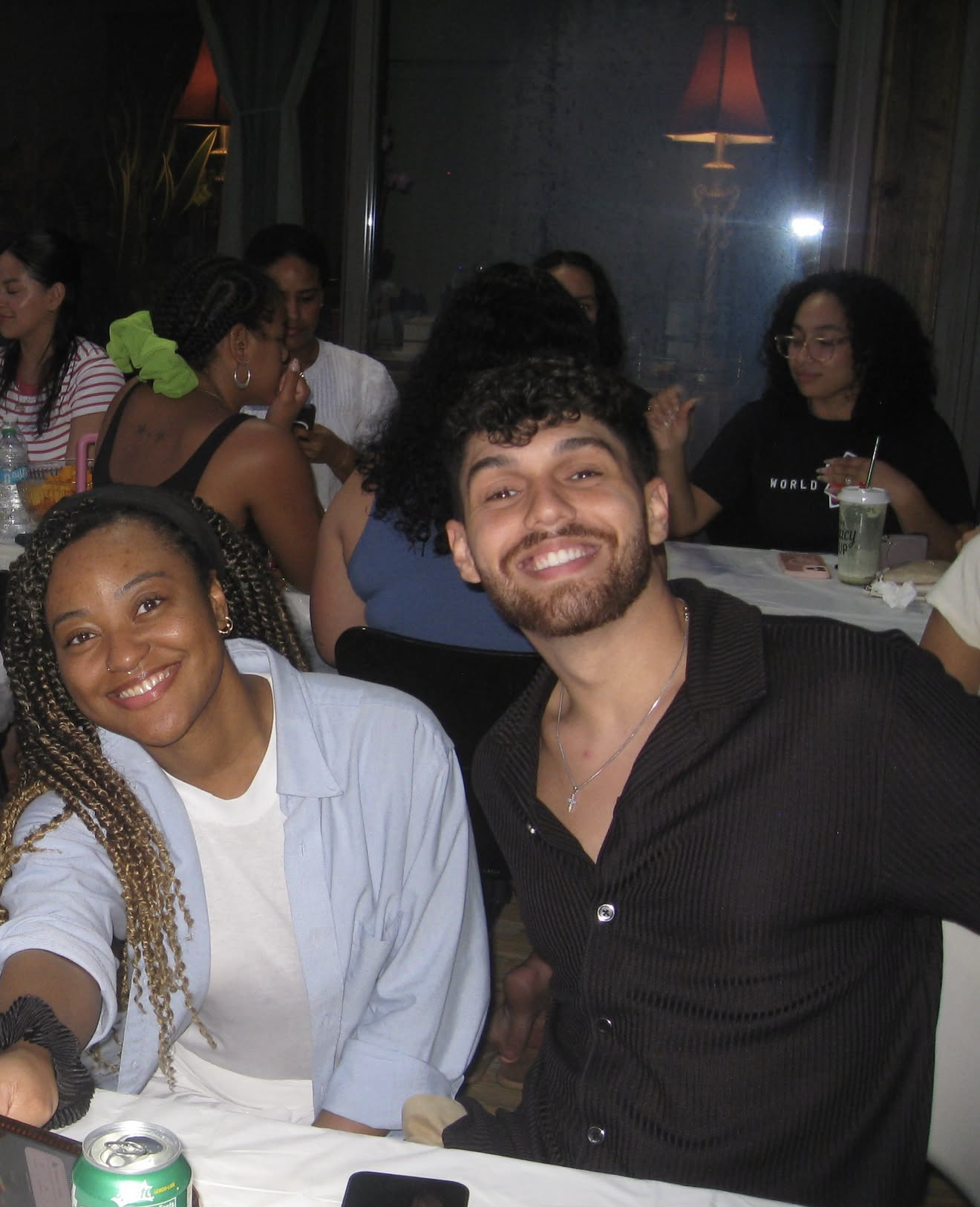 Smiling young woman with braided hair and a white shirt, smiling young man with curly hair and a black shirt, seated at a dinner table with drinks and electronics, surrounded by people in a dimly lit restaurant or banquet setting.
