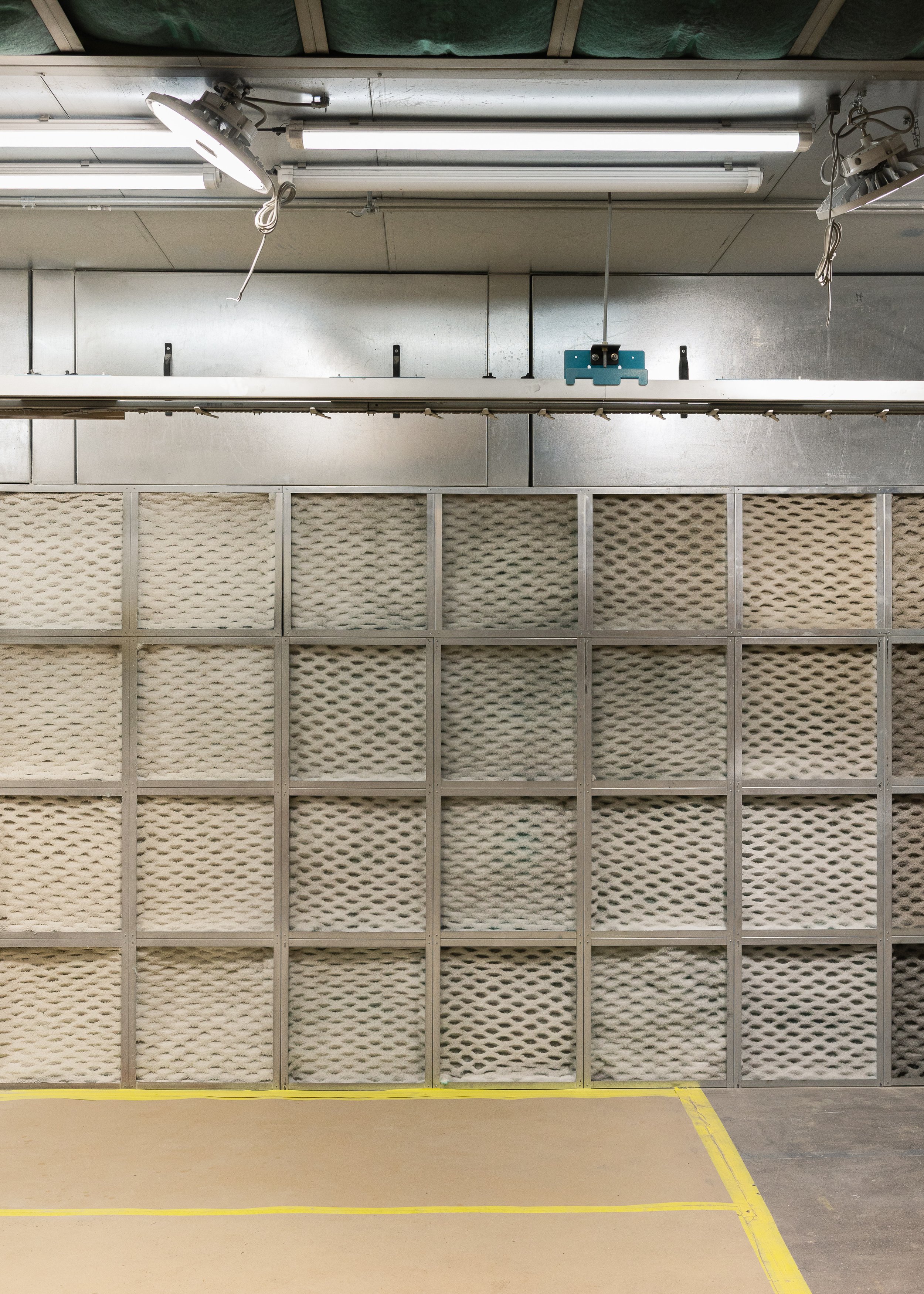 Industrial space with metal wall, fluorescent lights, and a grid of beige and gray acoustic panels. Yellow lines mark sections on the concrete floor.