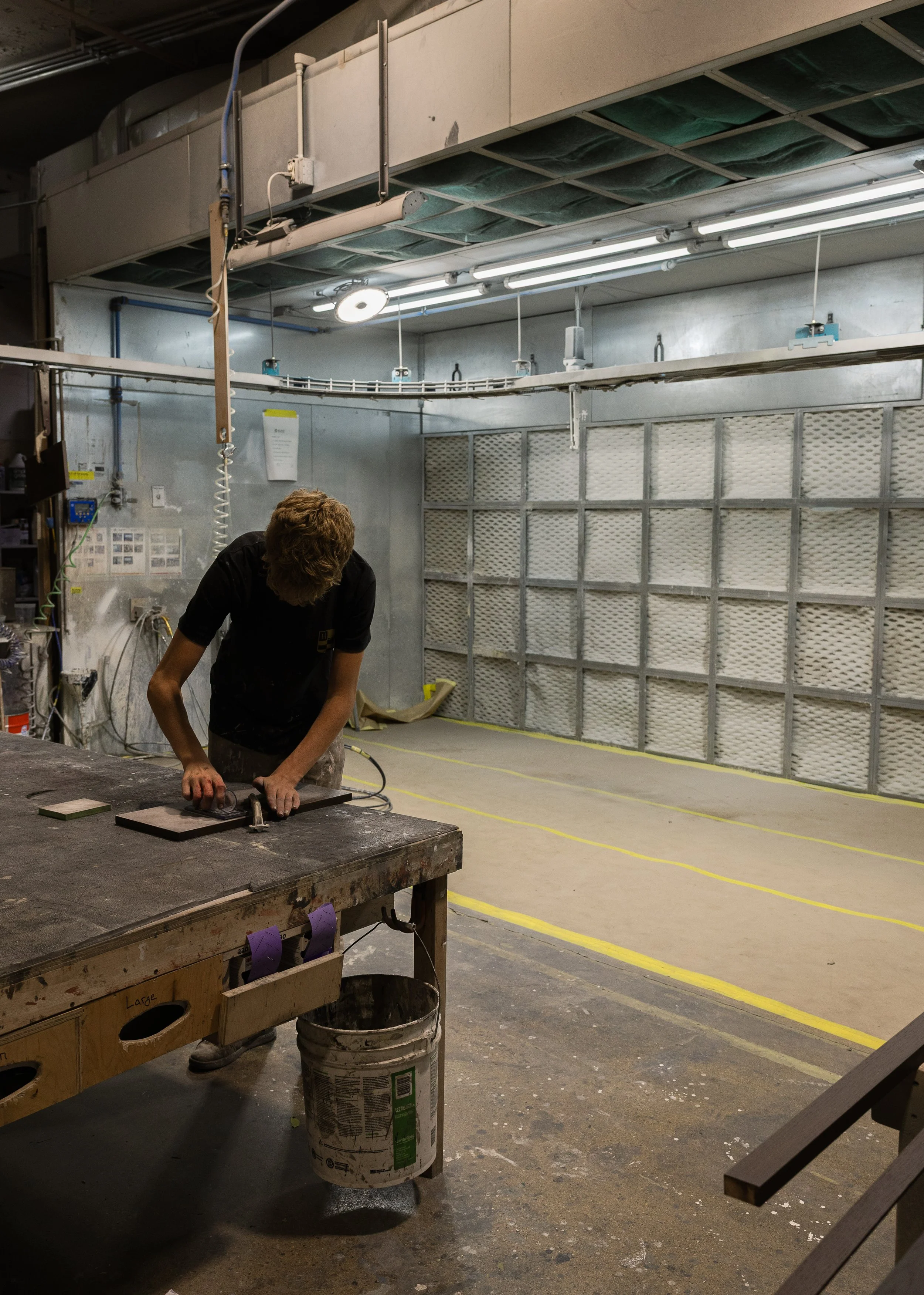 A person working in an industrial or workshop setting, leaning over a work table with tools, with spray booths or large filters on the back wall and overhead fluorescent lighting.