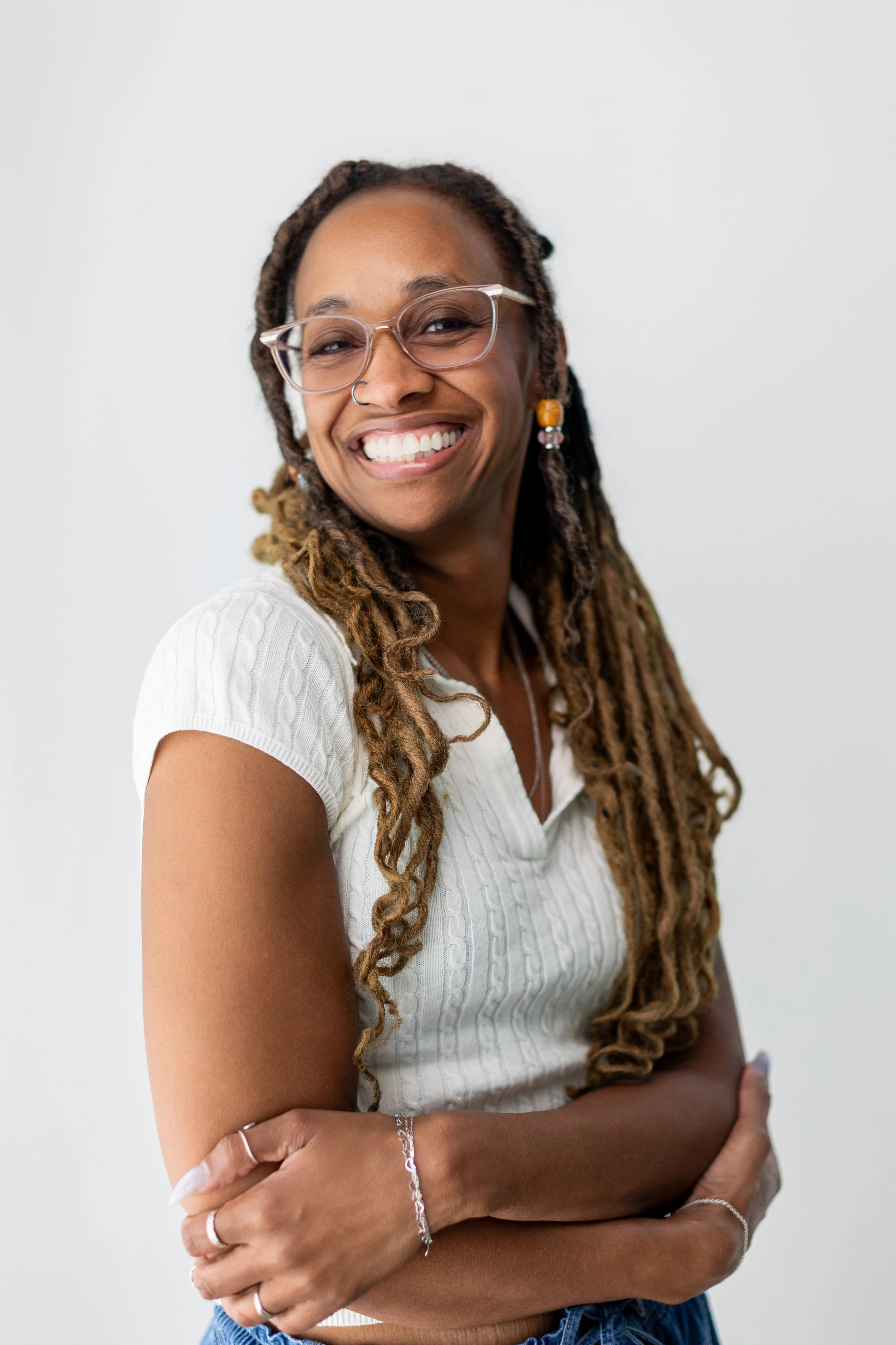 A woman with glasses, dreadlocks, and a nose ring smiling with arms crossed, wearing a white sweater and jewelry, against a plain white background.
