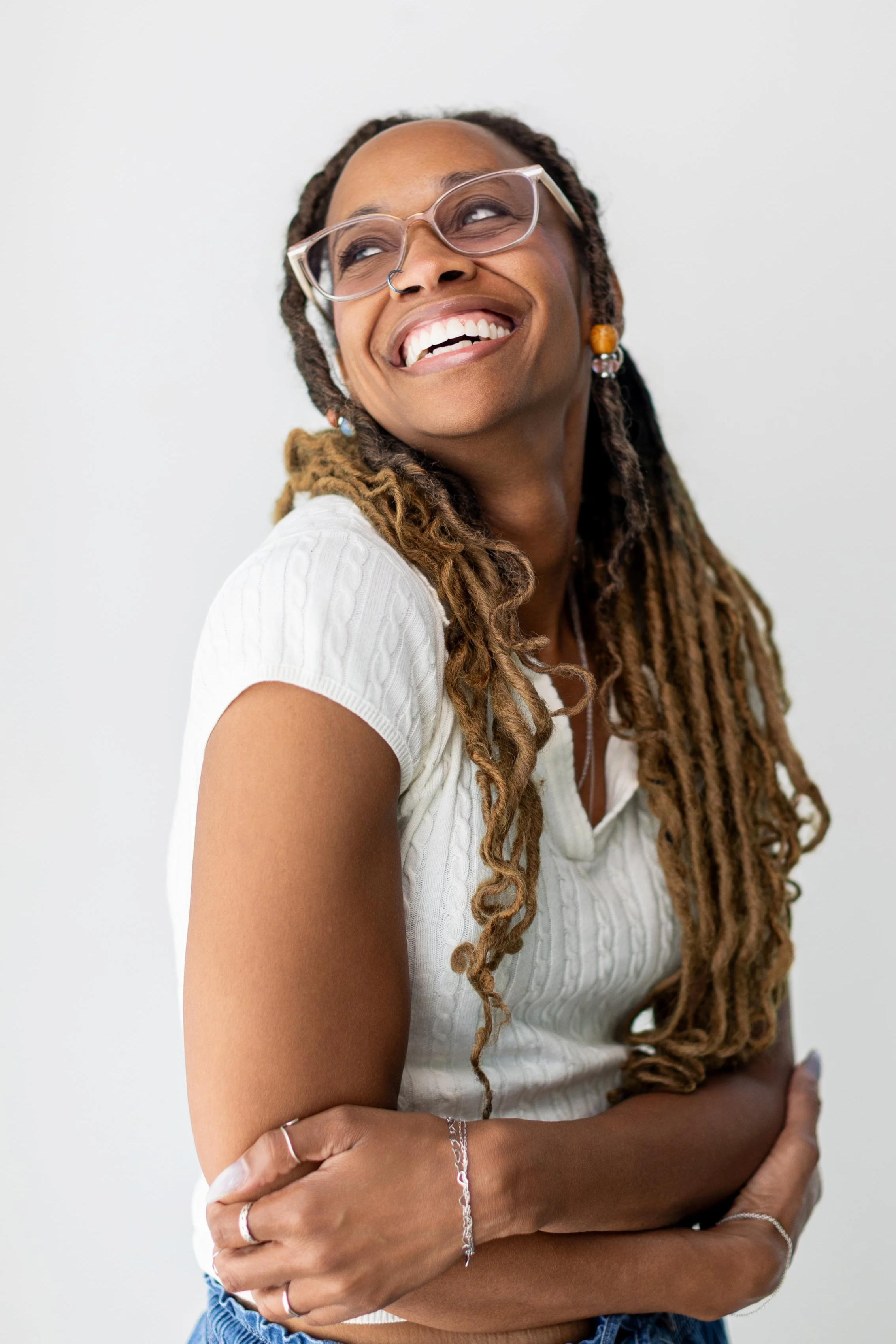 Portrait of a smiling woman with glasses, dreadlocks, and jewelry, wearing a white top, standing against a plain white background.