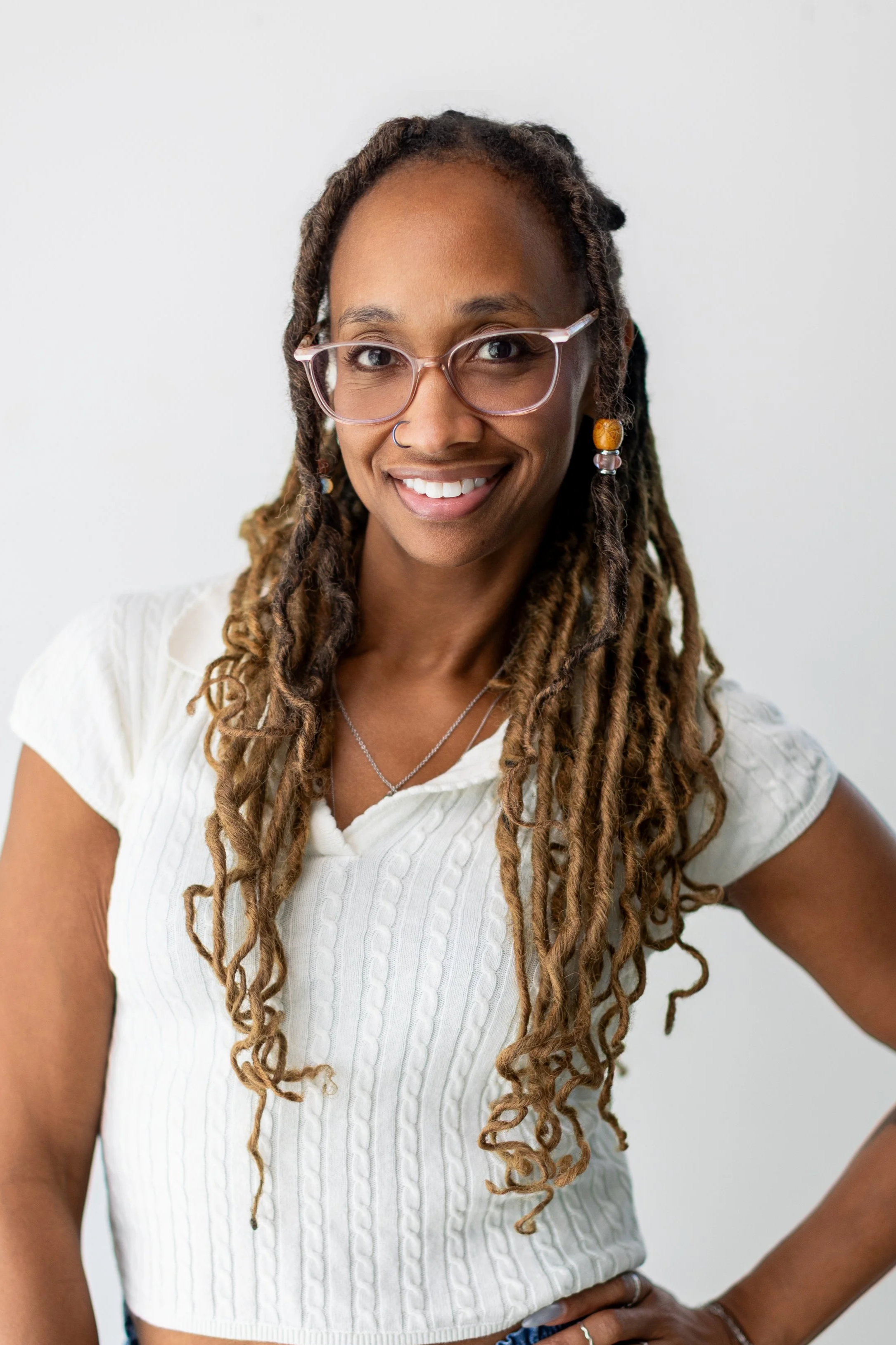 Close-up of a young woman with dreadlocks wearing lace glasses, jewelry, and a white cable-knit crop top, smiling against a plain light background.