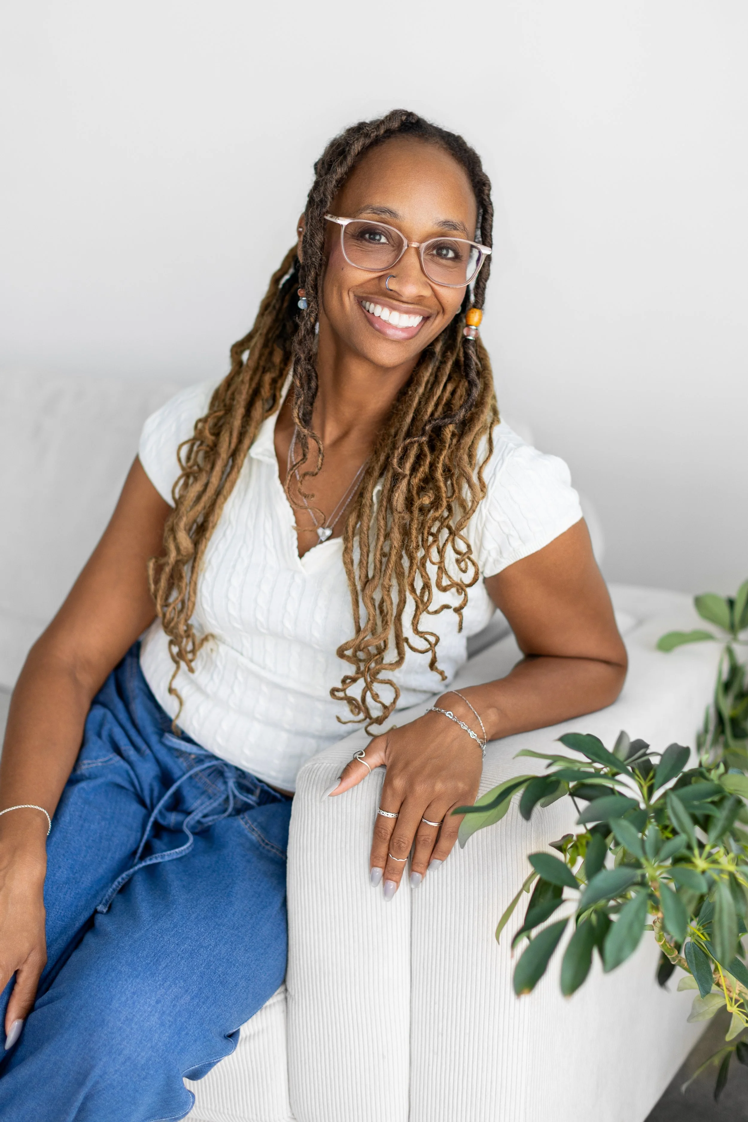 A woman with dreadlocks wearing glasses, a nose ring, and a white shirt, sitting on a white sofa, smiling at the camera with plants around her.