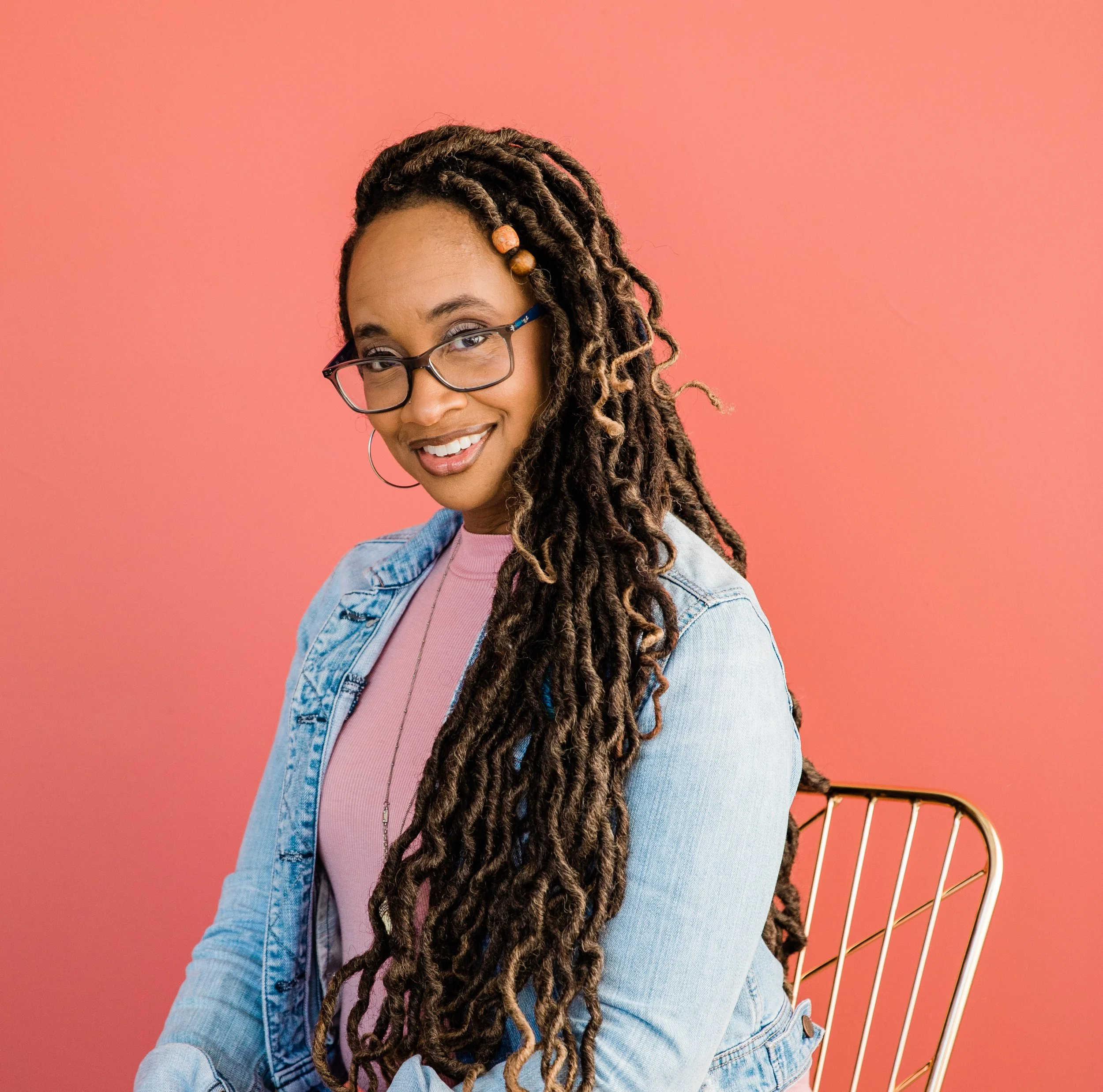 Smiling woman with glasses and dreadlocks wearing a pink top, denim jacket, and hoop earrings against a pink background with a gold and white chair.