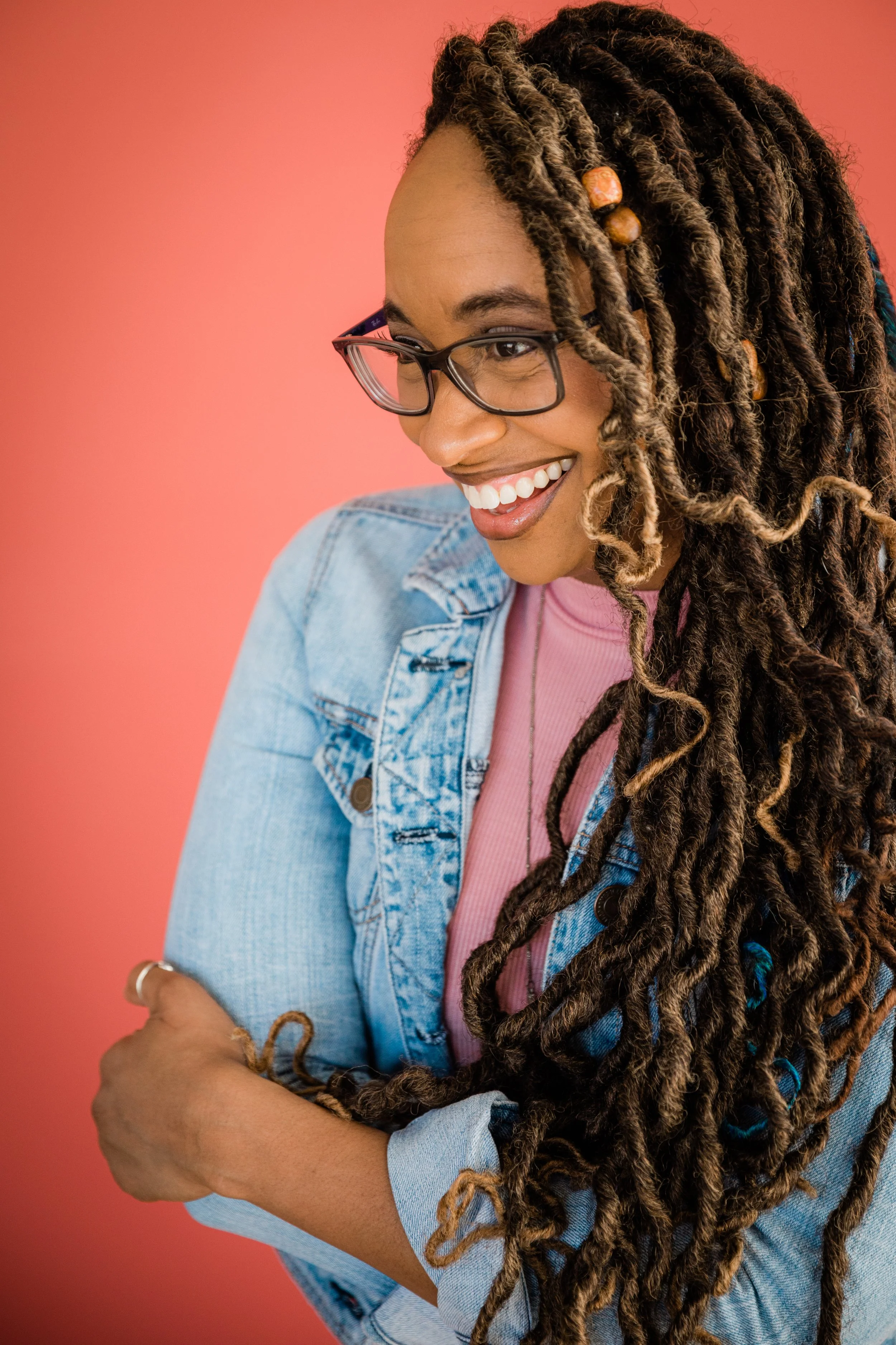 A smiling woman with glasses and long dreadlocks, wearing a denim jacket over a pink shirt, against a pink background.