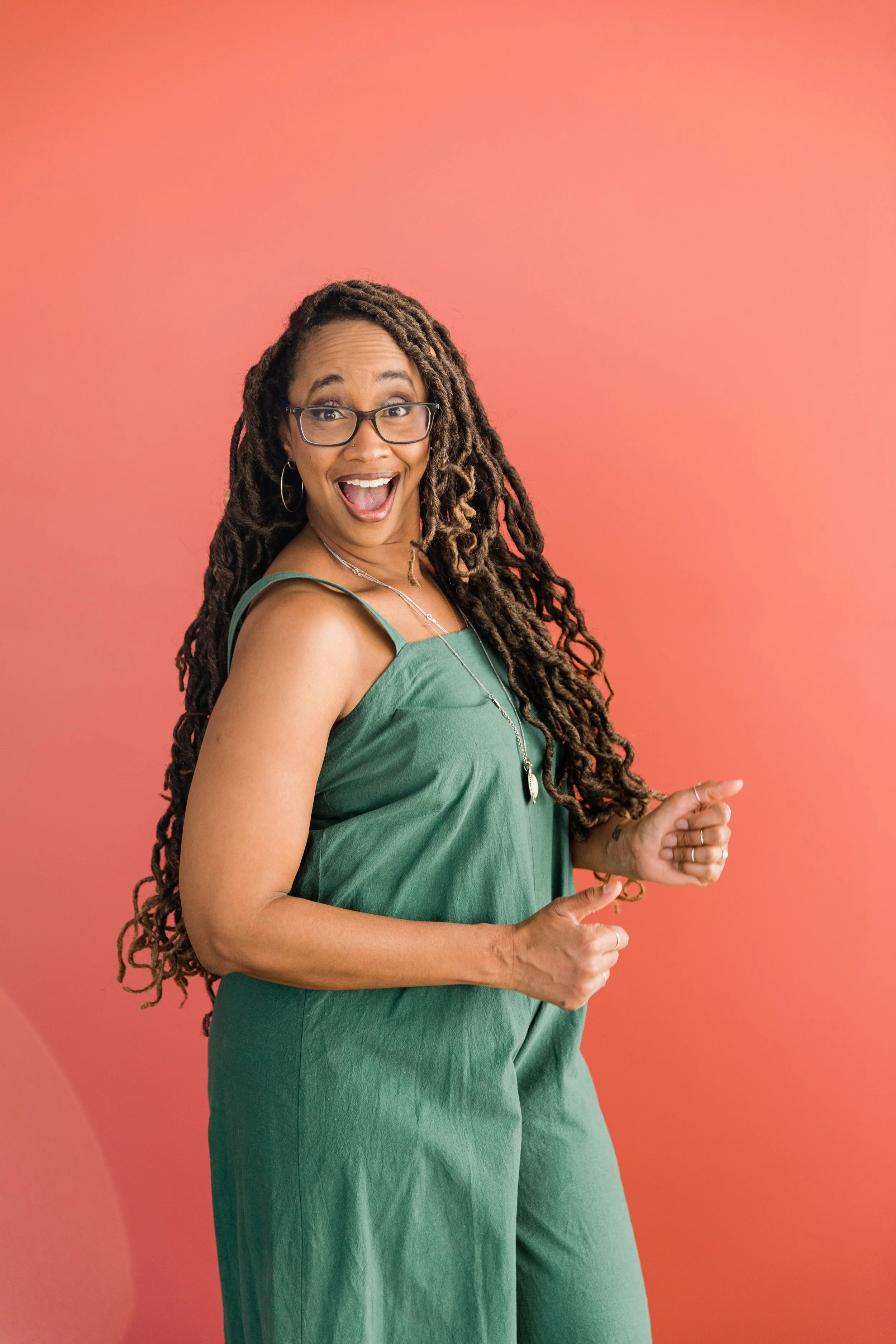 A woman with long curly hair, glasses, and hoop earrings, wearing a green dress, standing against a pink background, smiling enthusiastically with an open mouth and raised hands.