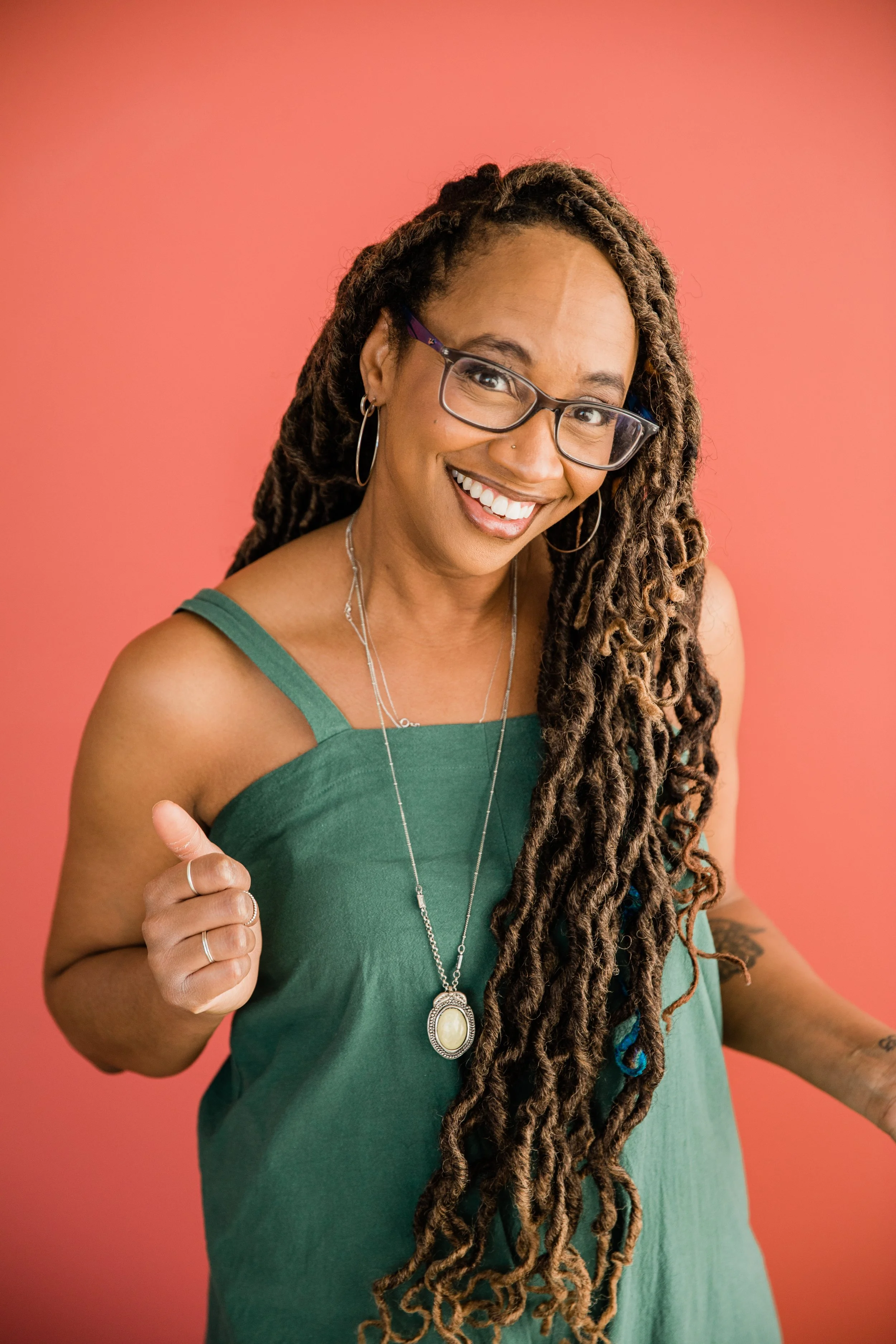 Smiling woman with long dreadlocks, wearing glasses, hoop earrings, and a necklace, giving a thumbs-up gesture against a pink background.