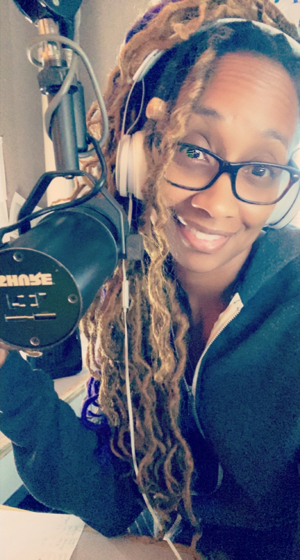 A woman with glasses and long, curly dreadlocks, smiling, wearing headphones and a black zip-up hoodie while sitting at a desk with a microphone in front of her.