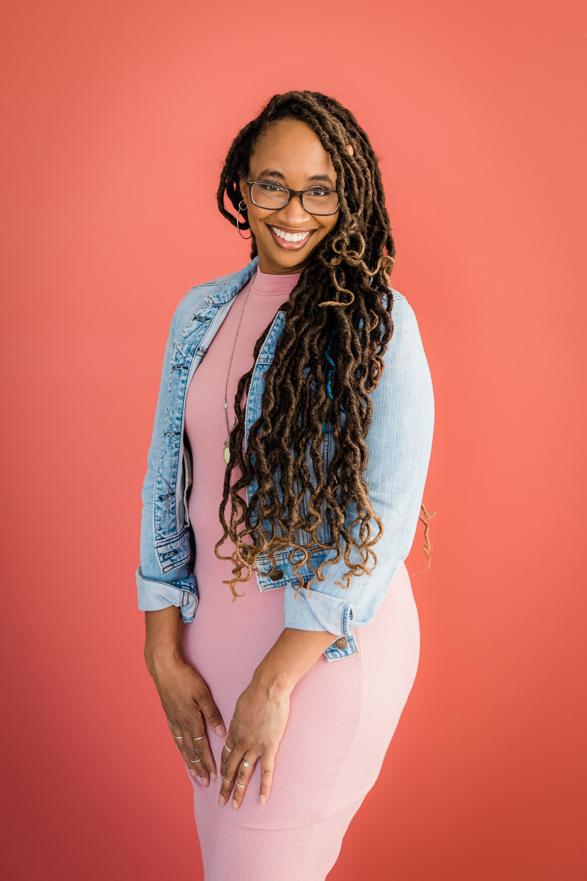 A woman with long curly hair, wearing glasses, a pink dress, and a denim jacket, standing against a pink background, smiling at the camera.