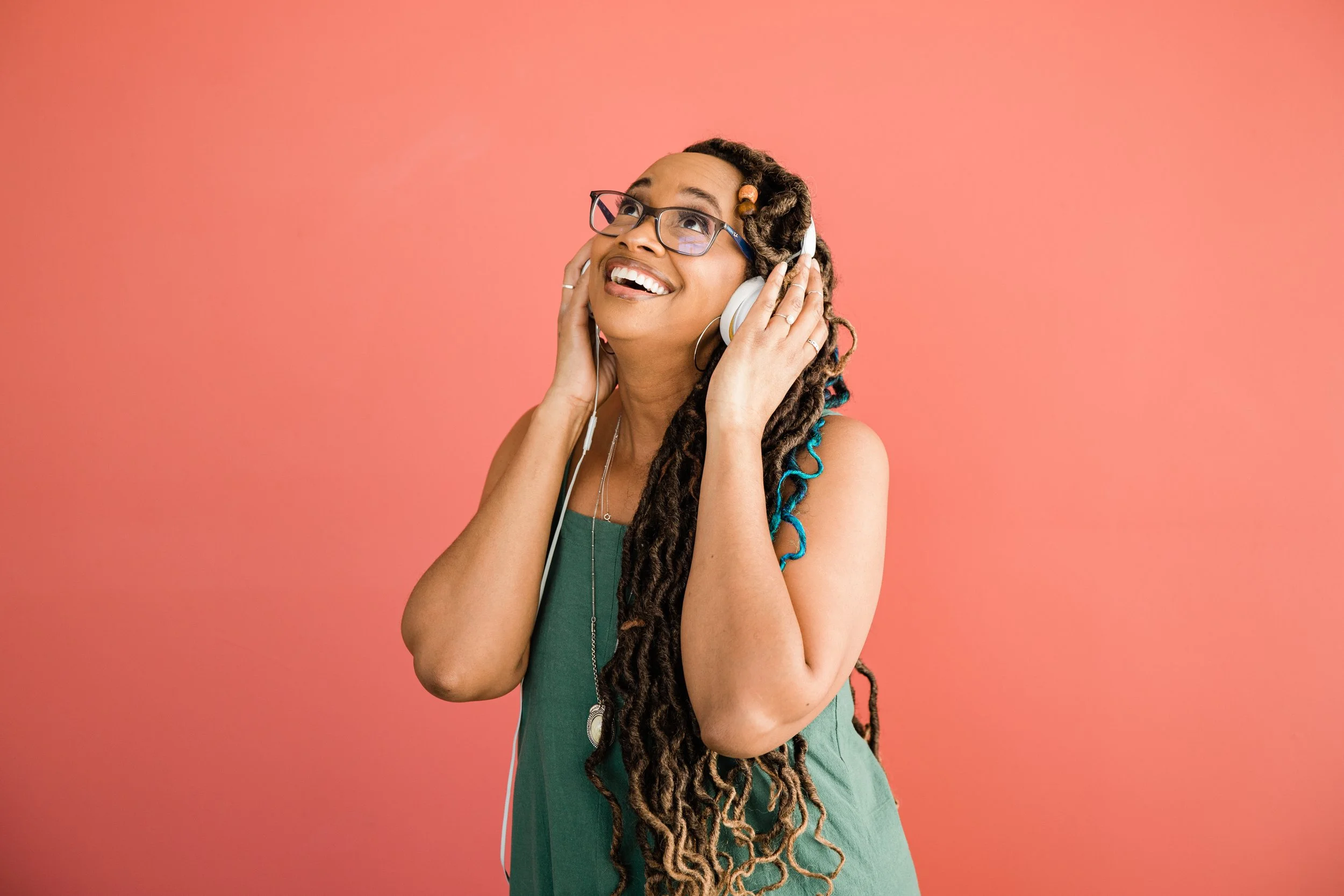 A woman with long dreadlocks wearing glasses and a green dress enjoys listening to music with headphones against a pink background.