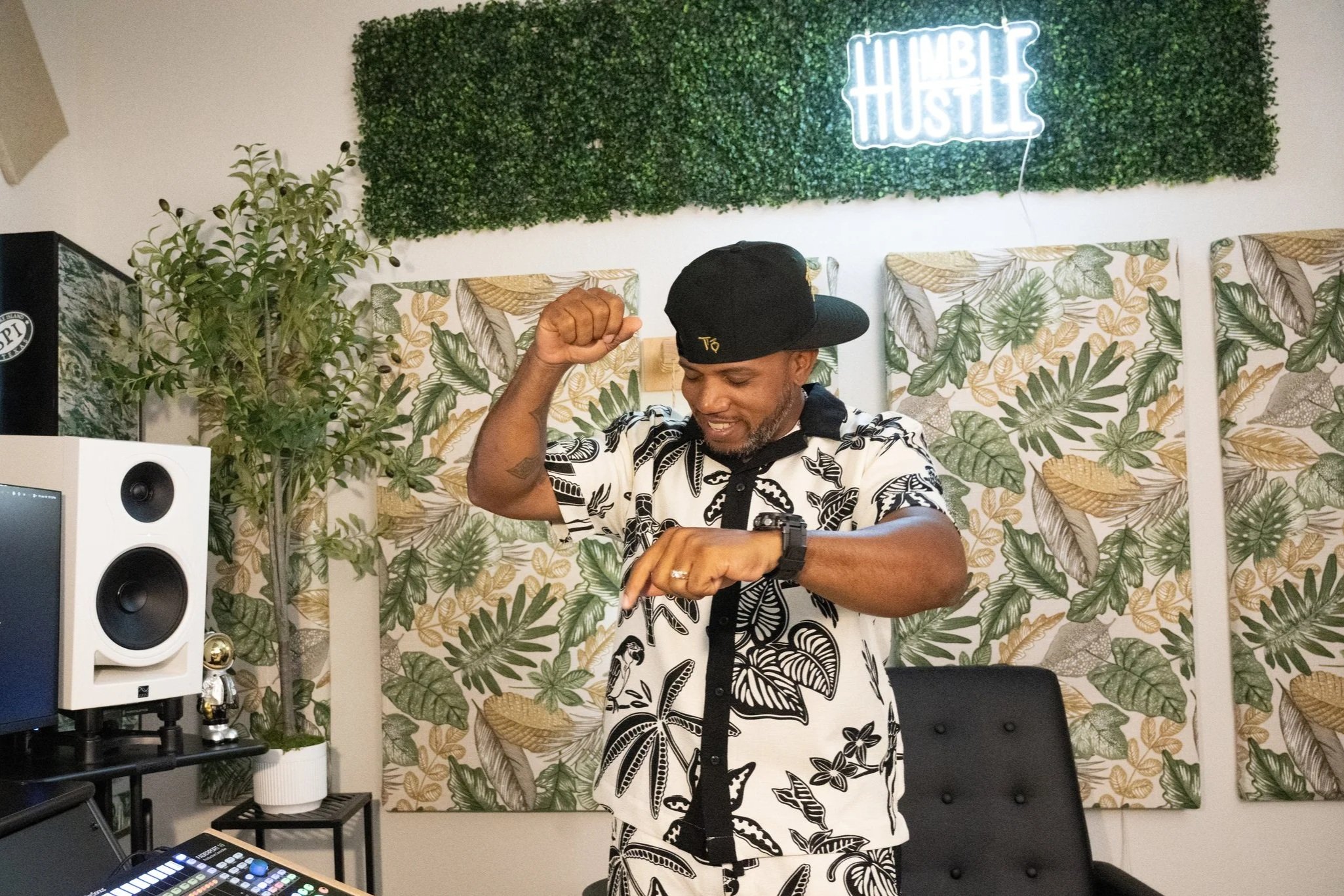A man wearing a patterned shirt and baseball cap, dancing or enjoying himself in a room decorated with tropical wallpaper and a green leafy wall with a neon sign that says 'HUSTLE'.