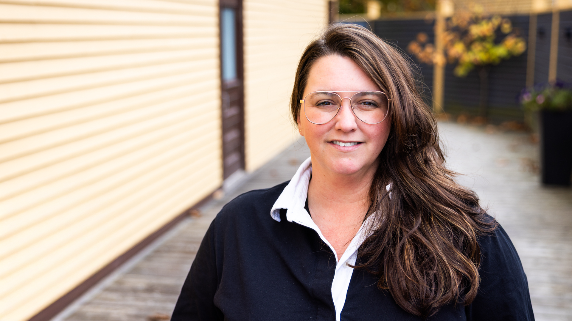 A woman with long brown hair wearing glasses, a black jacket, and a white collared shirt, smiling outdoors near a yellow wooden building with trees in the background.
