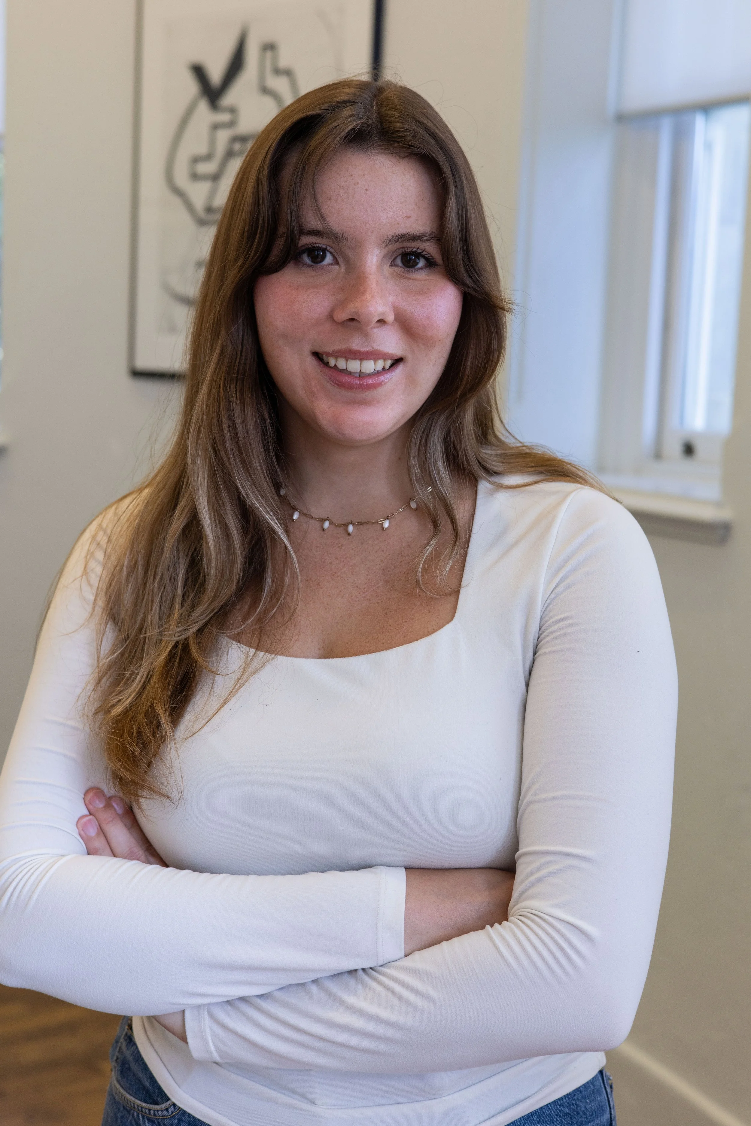 A woman with long, wavy brown hair, wearing a white long-sleeve shirt, standing with arms crossed, smiling in an indoor setting with a window and a wall art behind her.