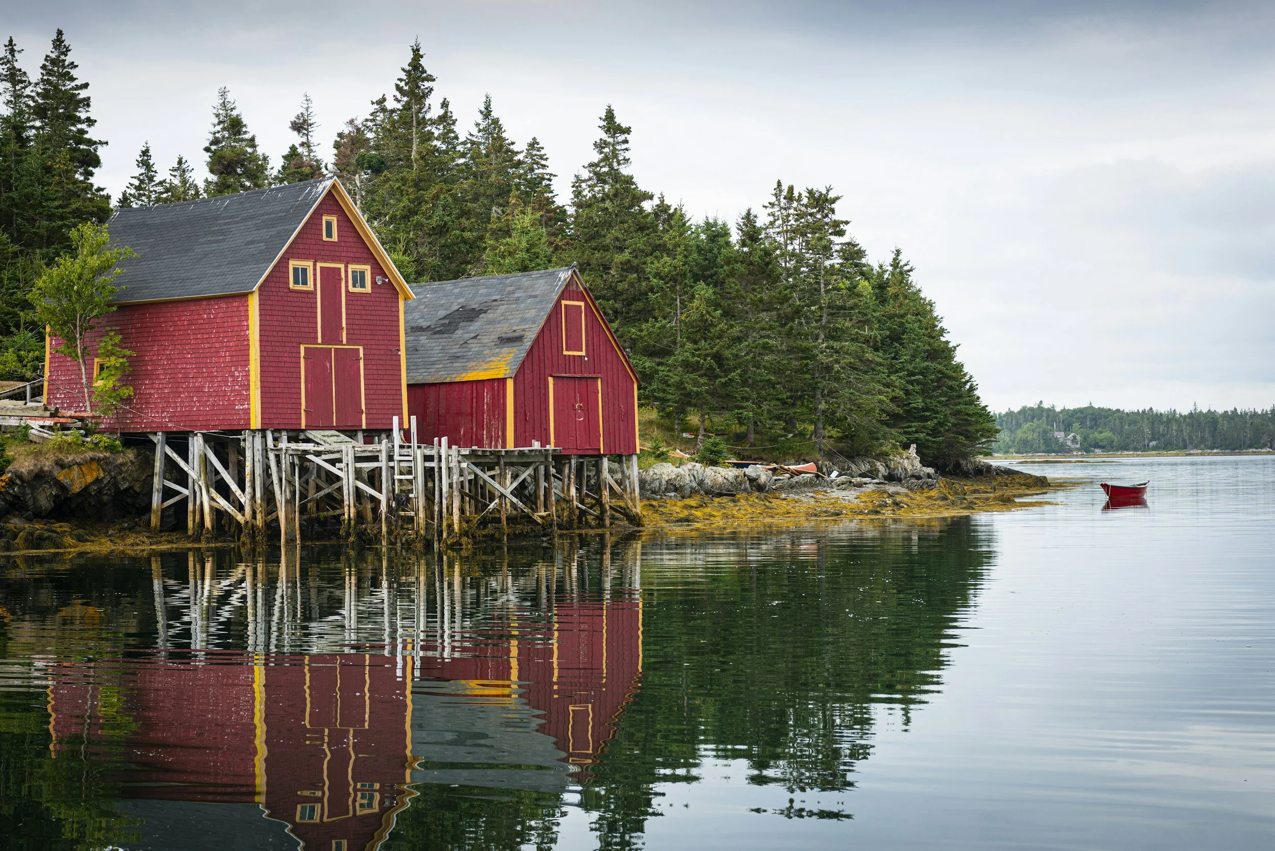 A red barn with yellow trim sits on wooden stilts by the water's edge, surrounded by evergreen trees under an overcast sky, reflecting in the calm water along a rocky shoreline.