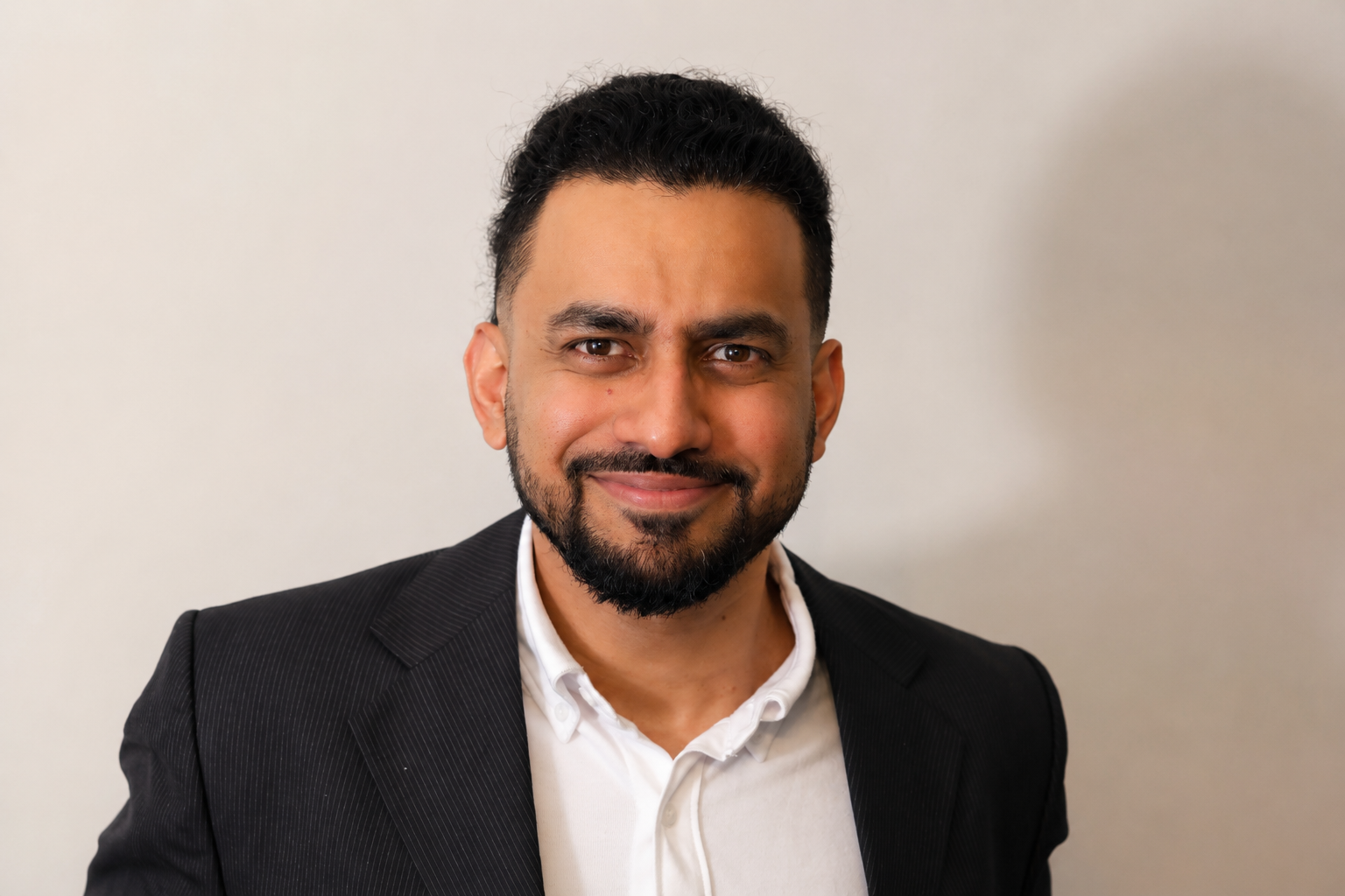 Professional man with dark hair and beard in a suit smiling at the camera against a plain wall.