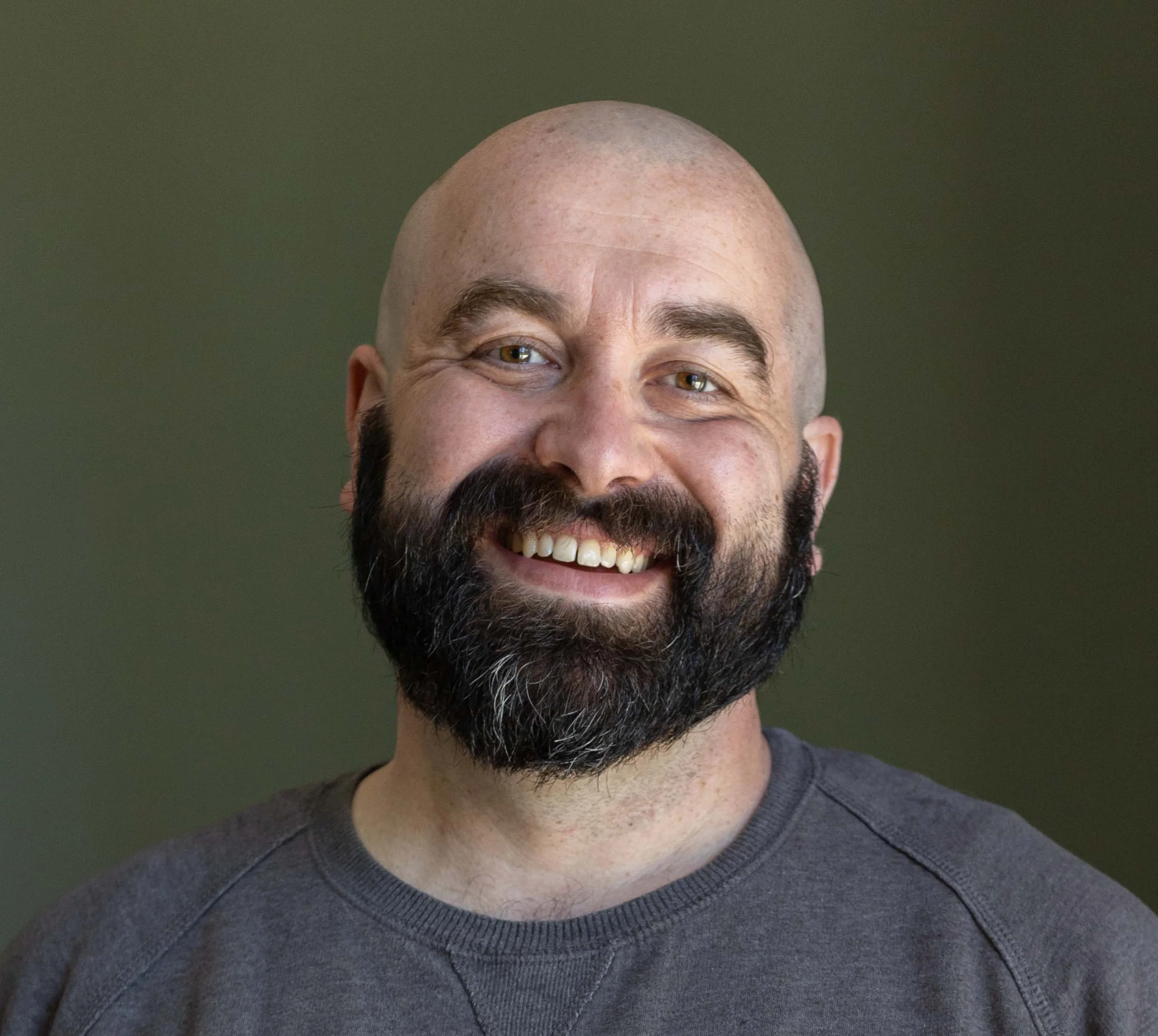 Close-up of a smiling man with a bald head, thick beard, and wearing a gray shirt.