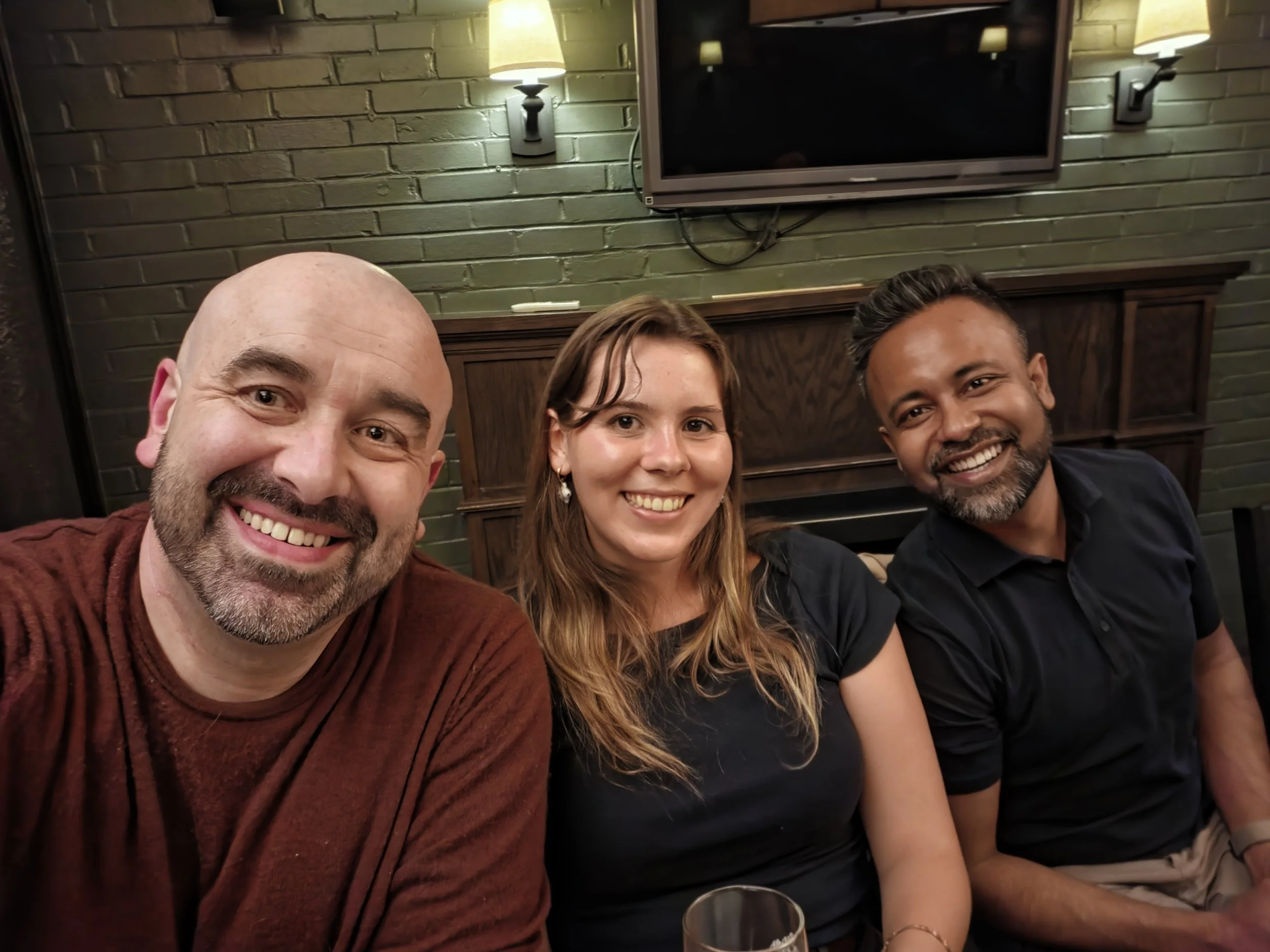 Close-up of three people smiling and taking a selfie in a room with a green brick wall and wall sconces.
