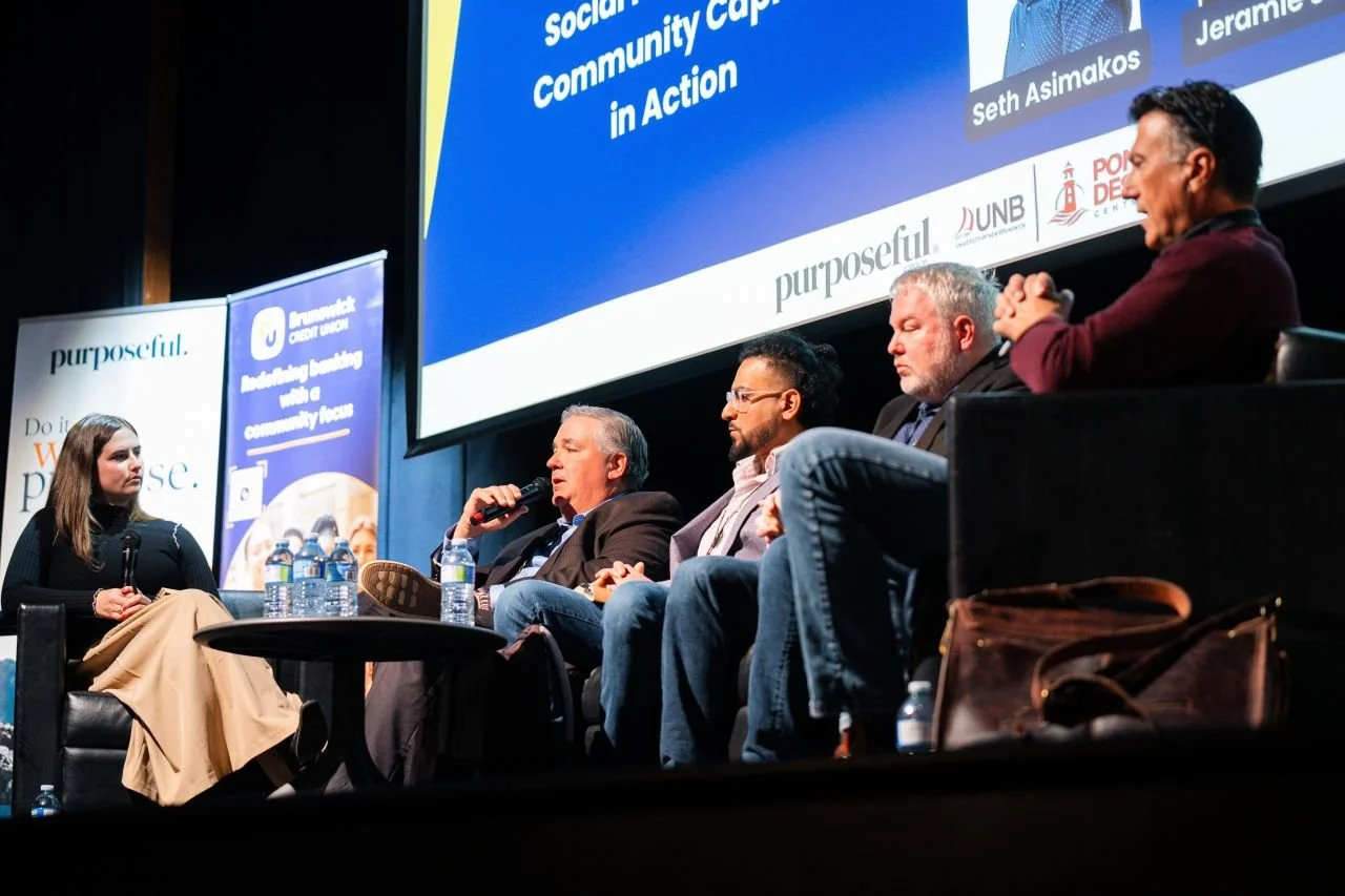 A panel discussion on stage with five people, including a woman and four men, seated in chairs; they are engaged in conversation with microphones and water bottles in front of them. A large screen displays event information behind them, with a banner on the left side.