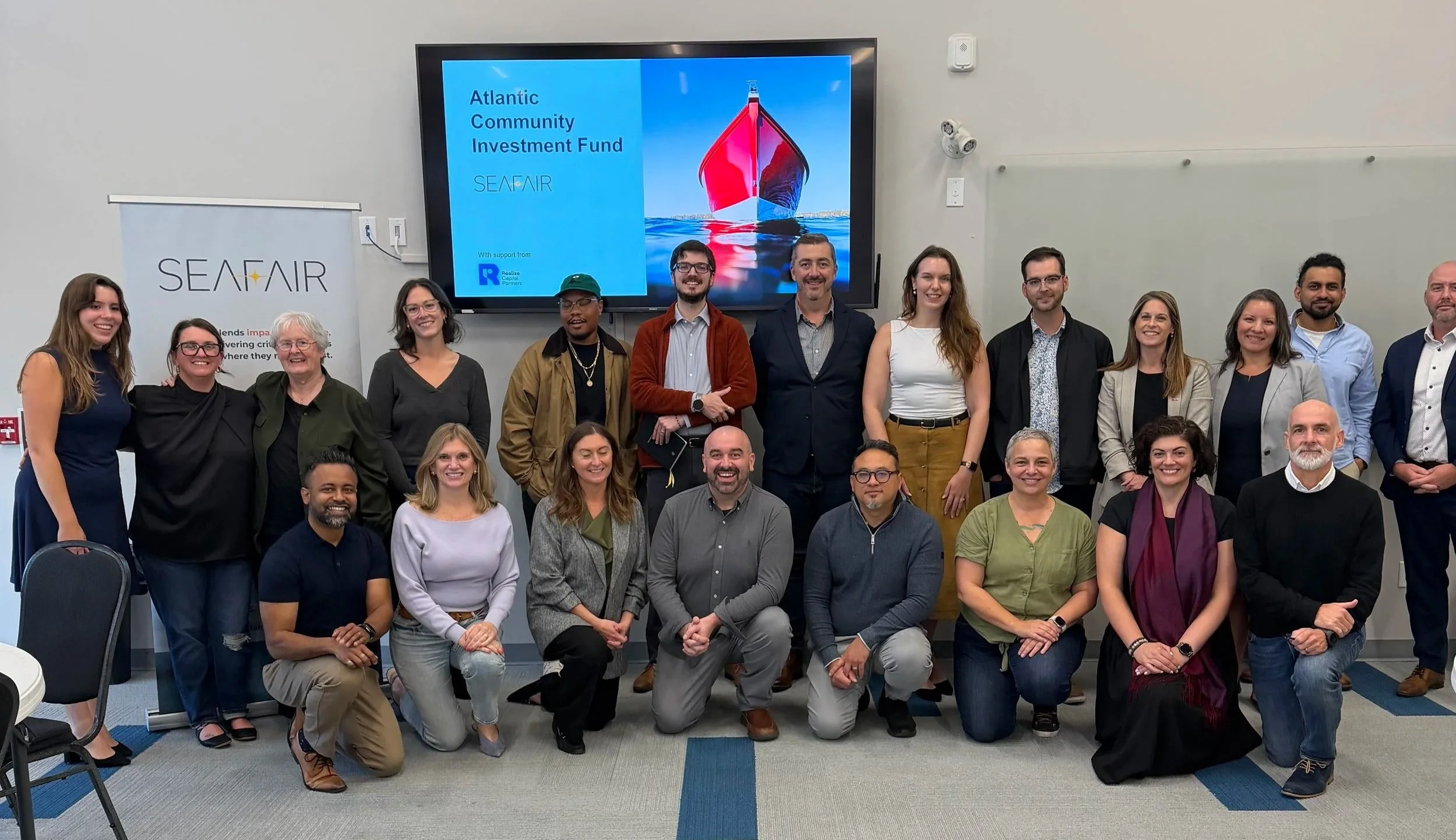 A group of diverse people posing for a photo in a conference room, with a large screen displaying 'Atlantic Community Investment Fund' and a boat image behind them.