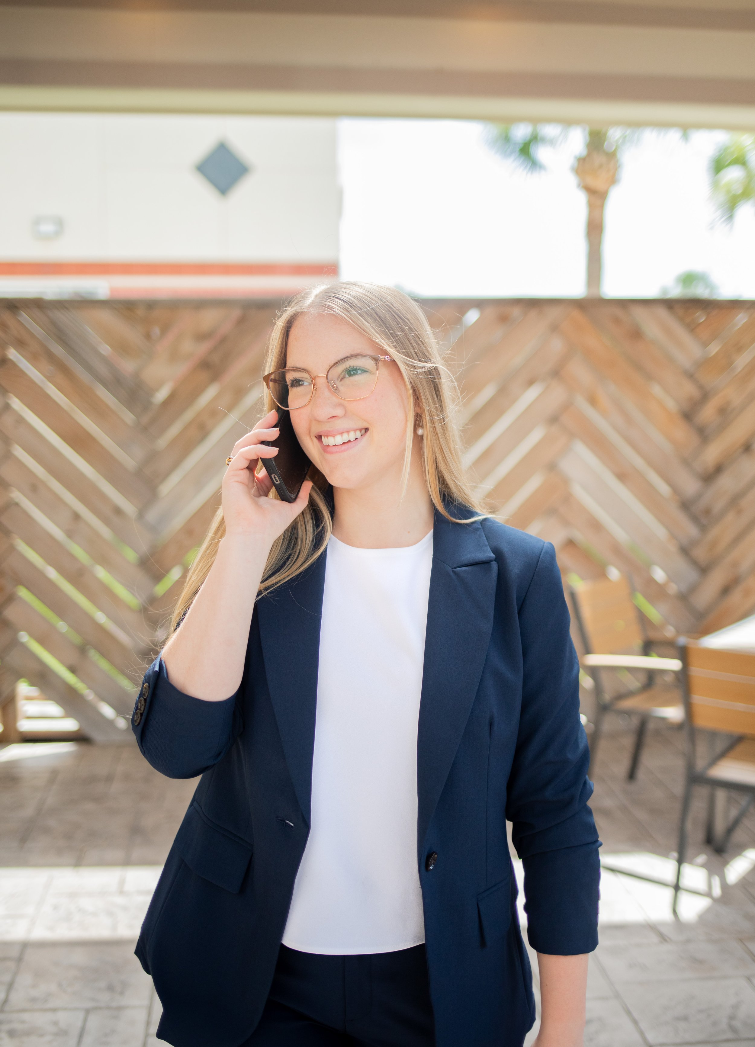 A woman with long blonde hair, glasses, and wearing a navy blue blazer, holding a phone to her ear and smiling, standing outdoors near a wooden fence with an umbrella and trees in the background.